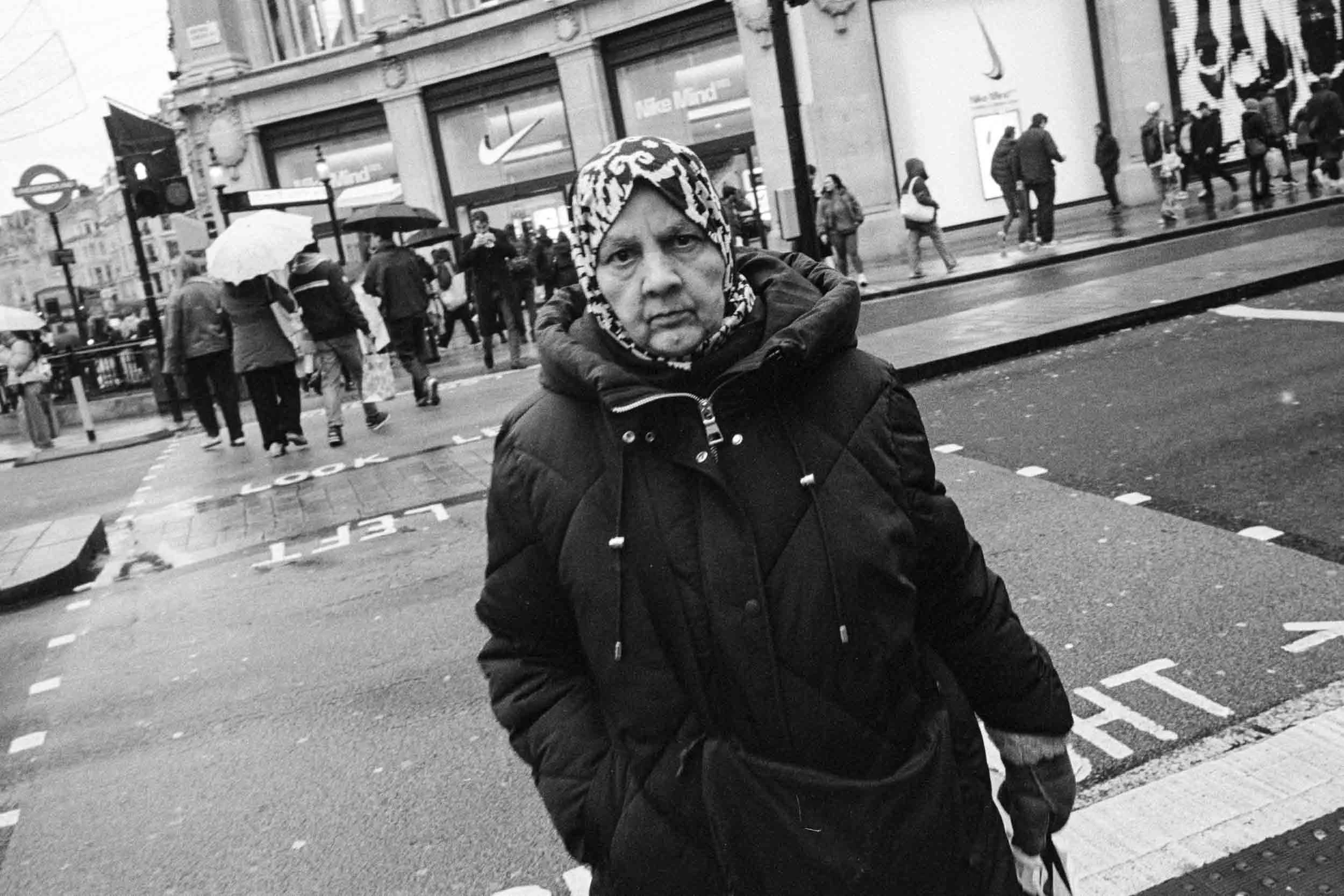 A woman wearing a patterned headscarf and a puffy winter coat standing on a busy city street in rain, with people walking with umbrellas in Oxford Circus London.