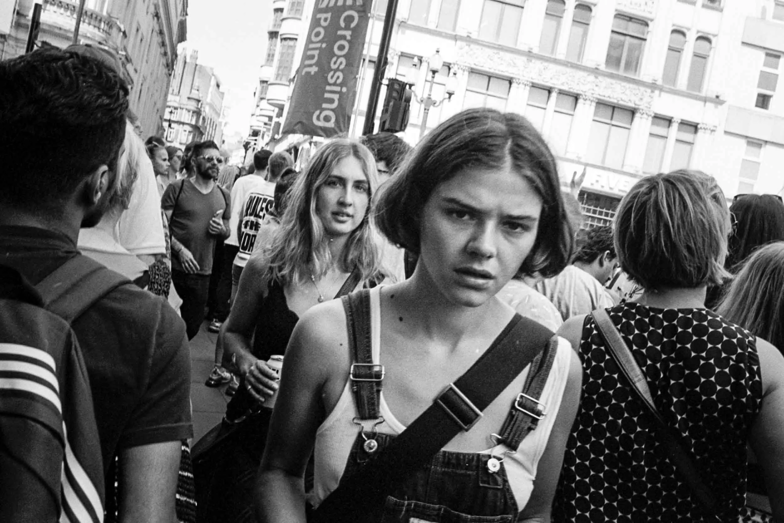 Crowd of people walking on a city street, with tall buildings in the background. Black and white photo.