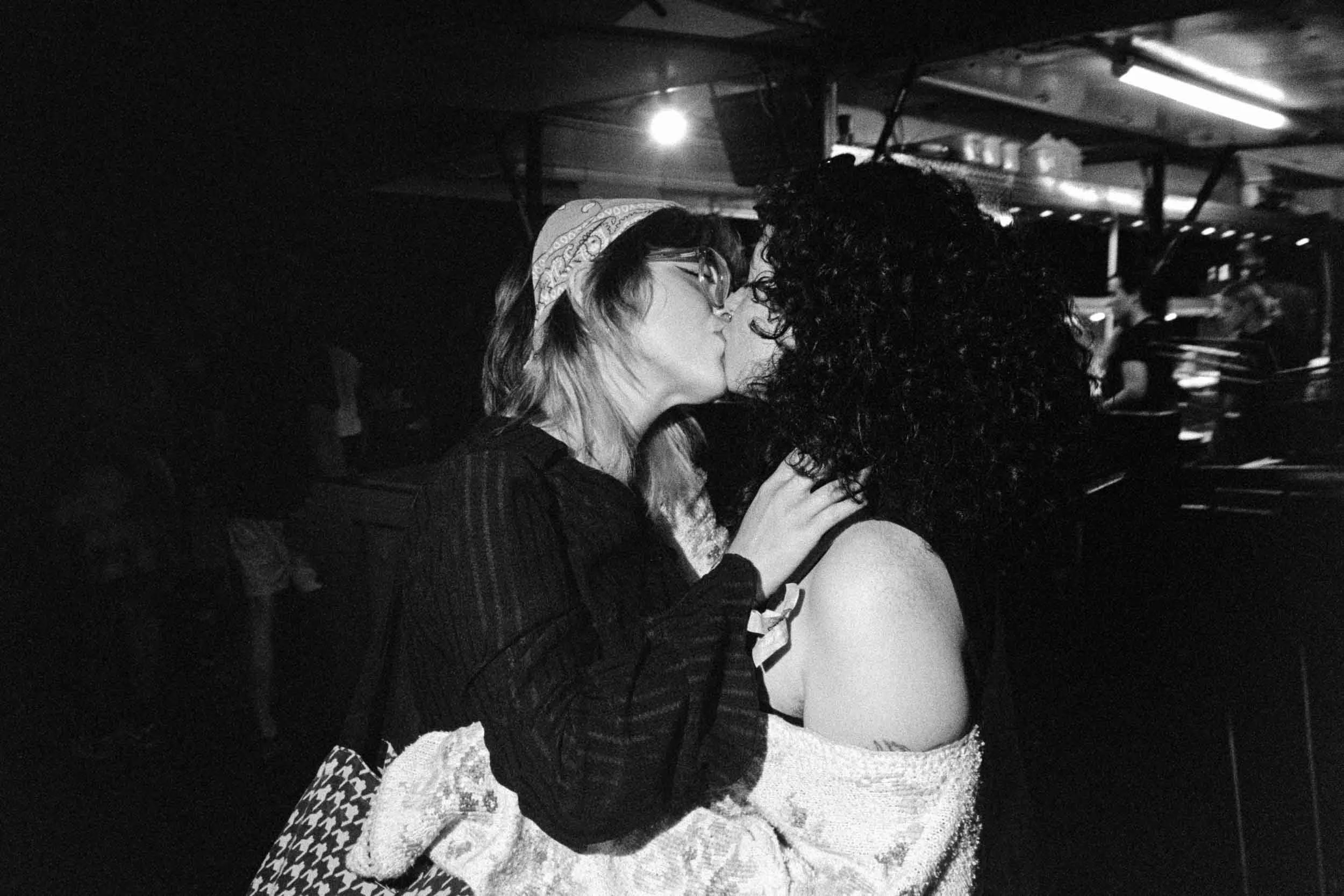 Two women sharing a kiss in a dimly lit environment, one wearing glasses and a bandana, the other with curly hair, with a person in the background in Canal Street at Manchester Pride.