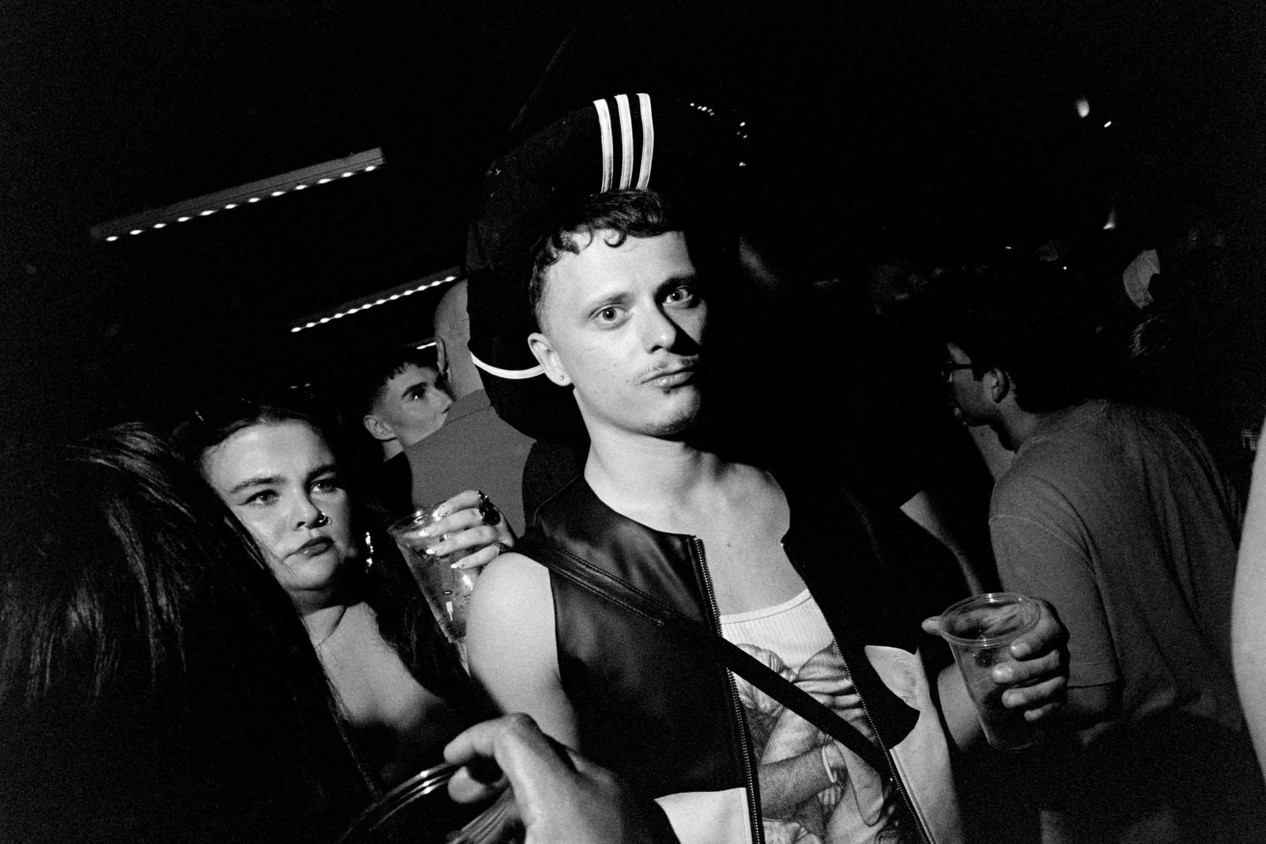 Black and white photo of a young man with curly hair and earrings, holding a drink, surrounded by other people at a social gathering or party.