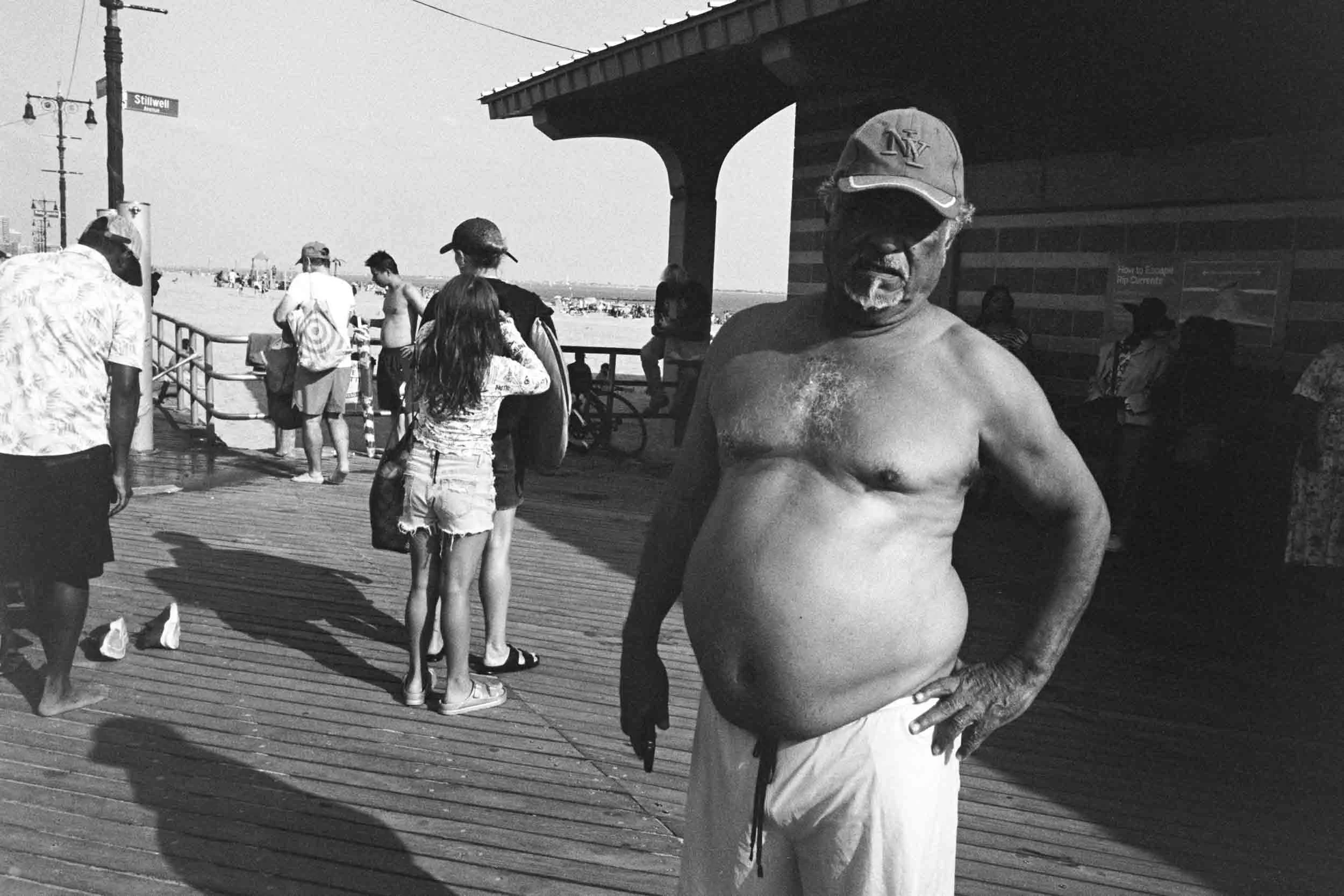 Man with a shirtless upper body wearing a cap, standing with hand on hip on a pier, with other people near beach and some structures in the background.