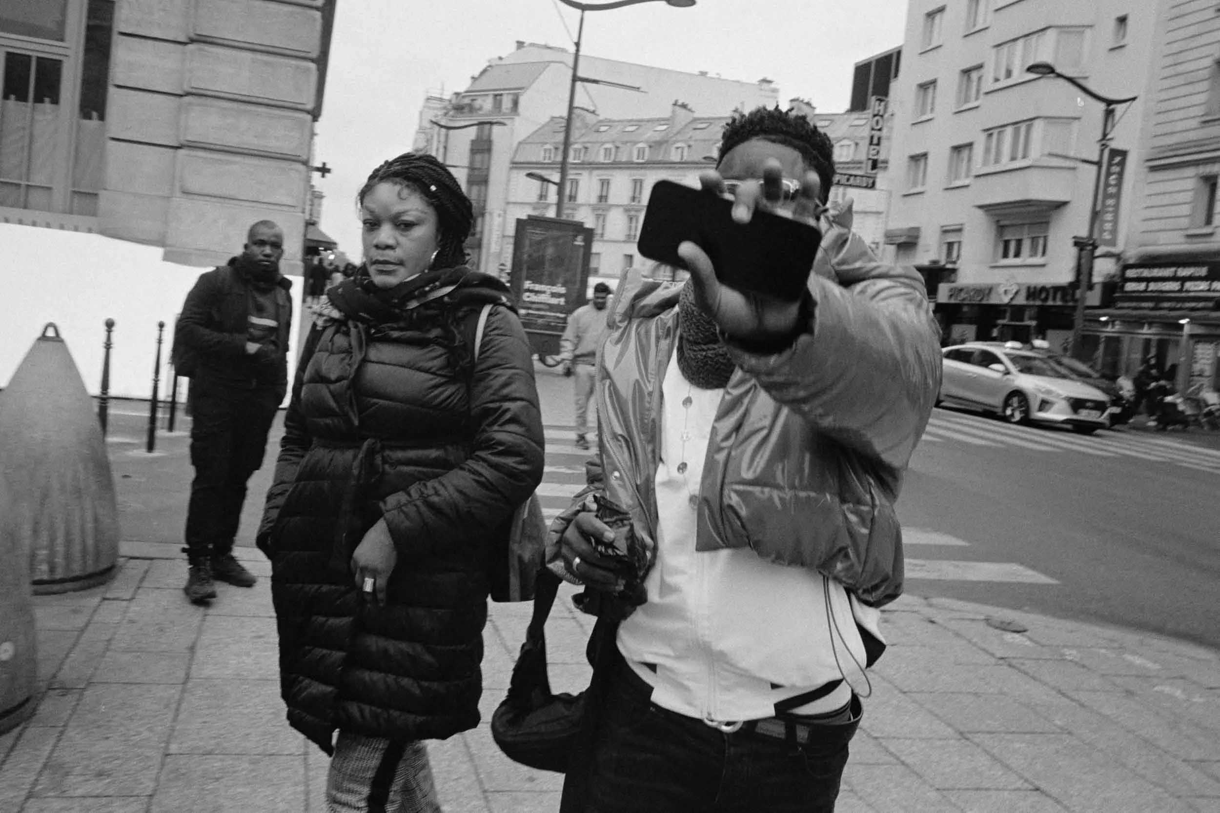 A black and white photograph of a city street with four people in the foreground. One woman with braided hair and a puffy jacket is looking to the side, another man with a hoodie and puffy jacket is pointing a phone toward the camera, and two other m
