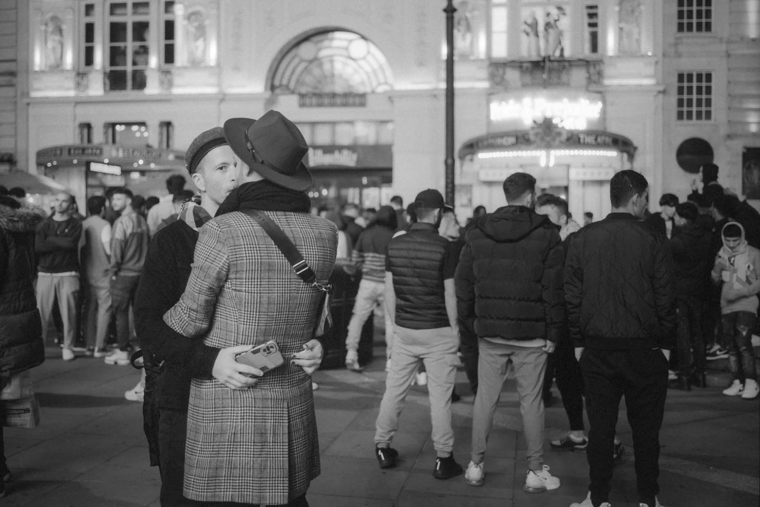 People gathered in a city street at night, with some wearing masks and hats, and buildings with lit windows in the background.