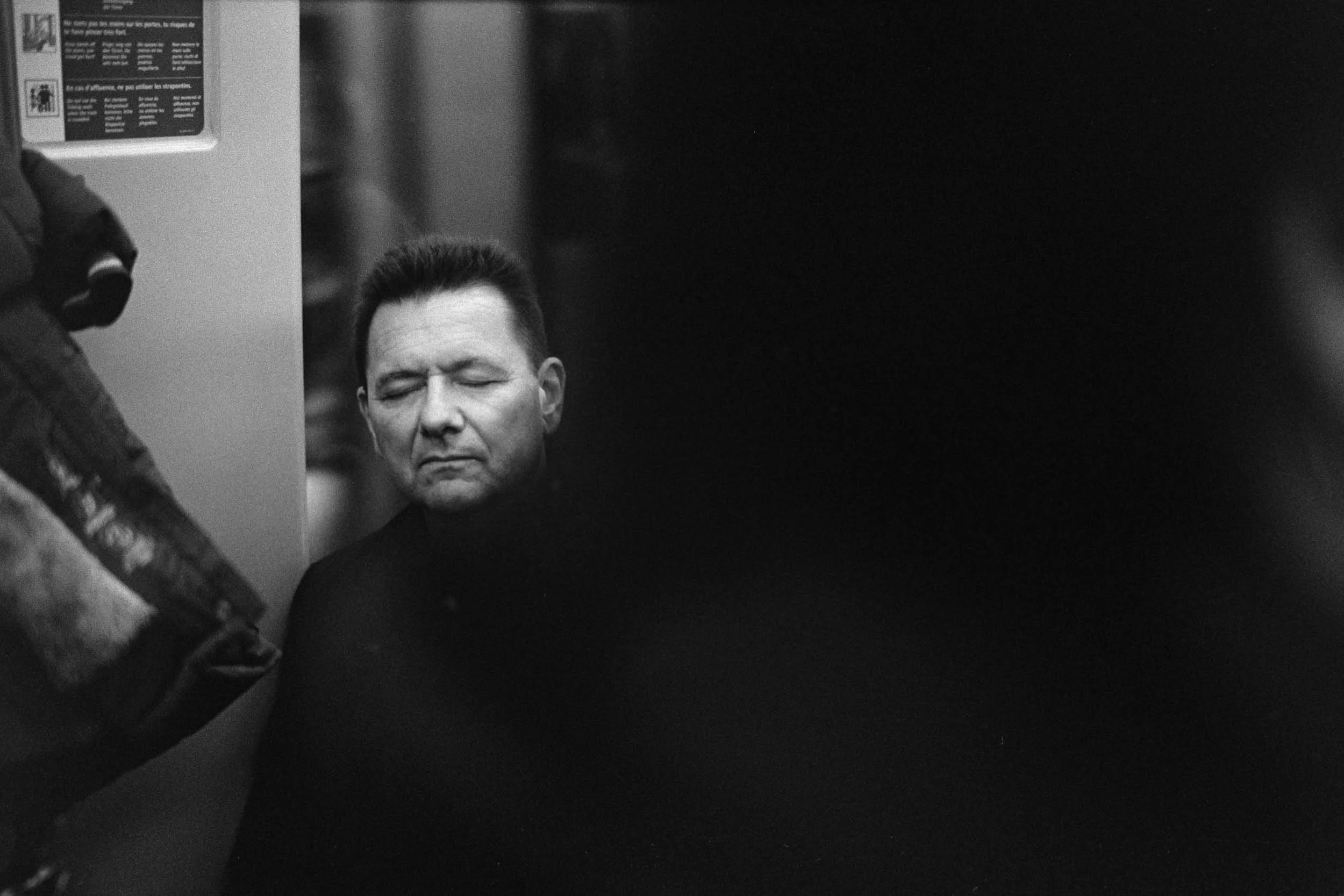Black and white photo of a man with closed eyes and slicked-back hair, with a mysterious expression, standing behind a dark surface.