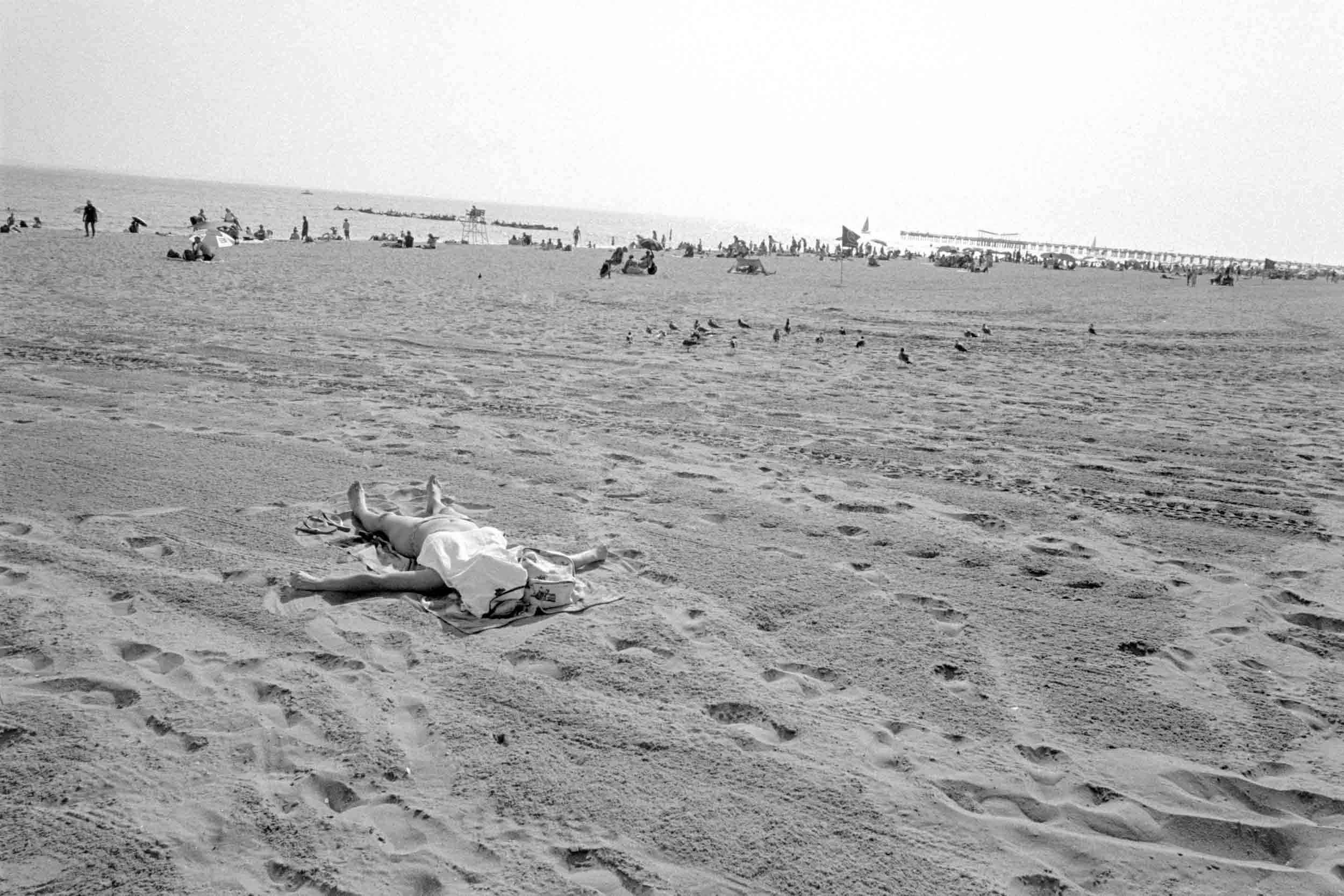Black and white photo of the beach at Coney Island with people lounging and walking along the sand, some under umbrellas and some near the water, with a pier in the background.