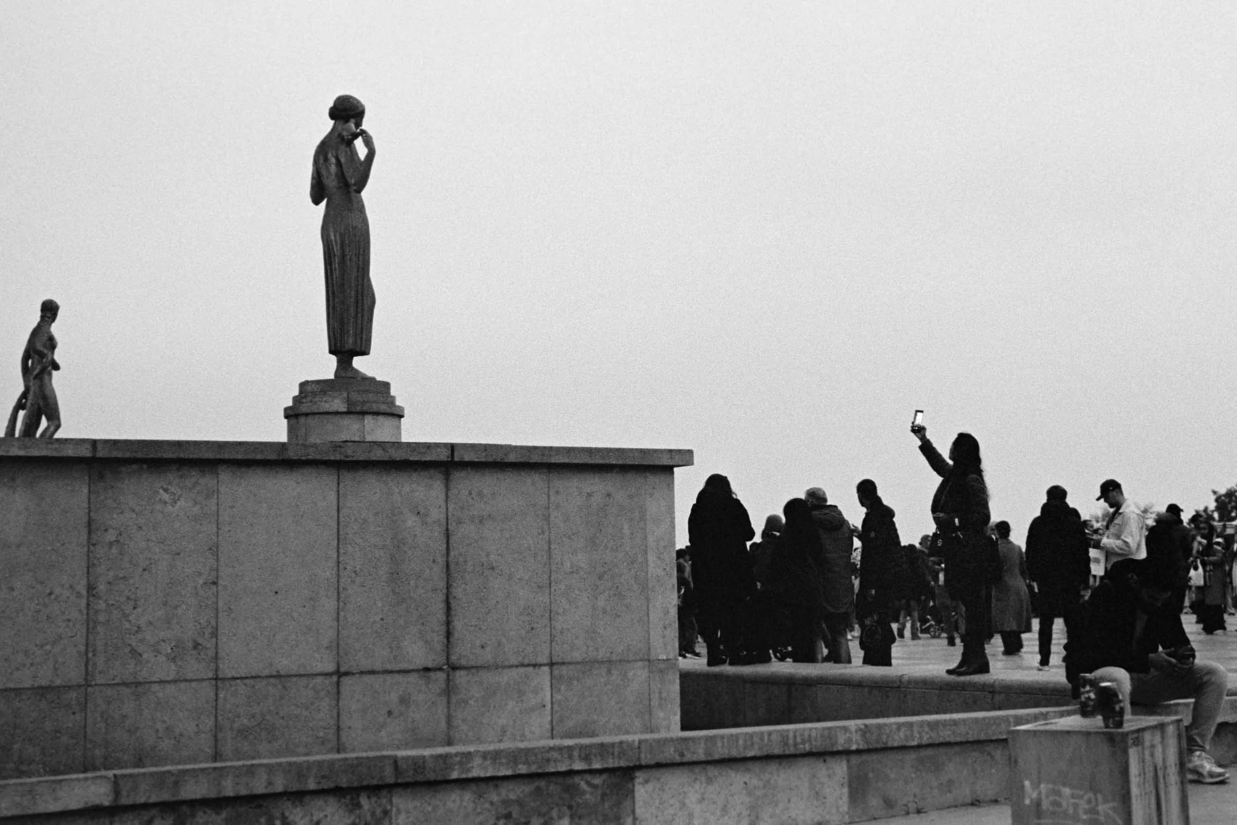 A black and white photo of a public square with a group of people and three statues on a raised platform. The statues depict women in traditional attire.