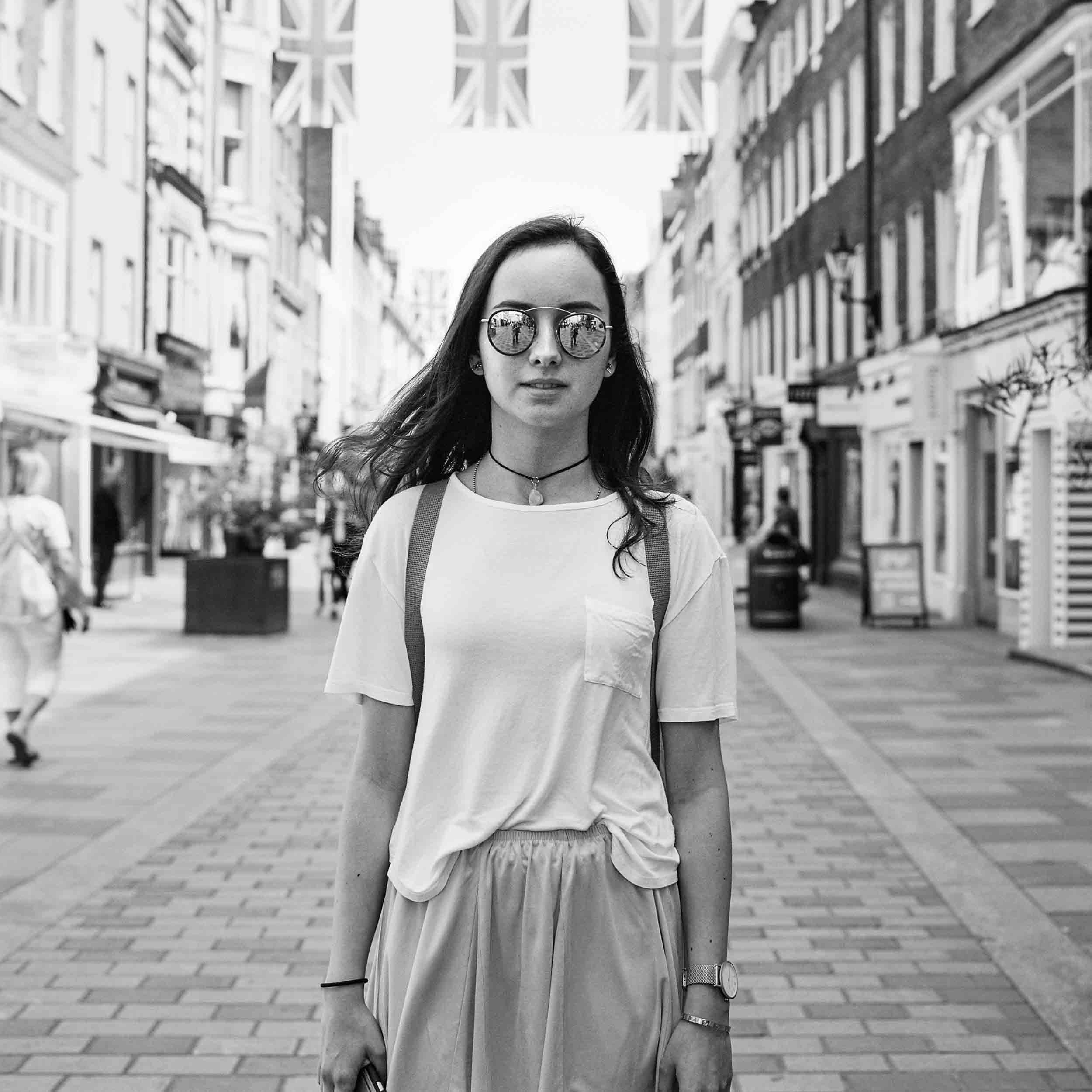 Black and white photograph of a young woman with long hair and sunglasses walking on a city street with tall buildings on either side.