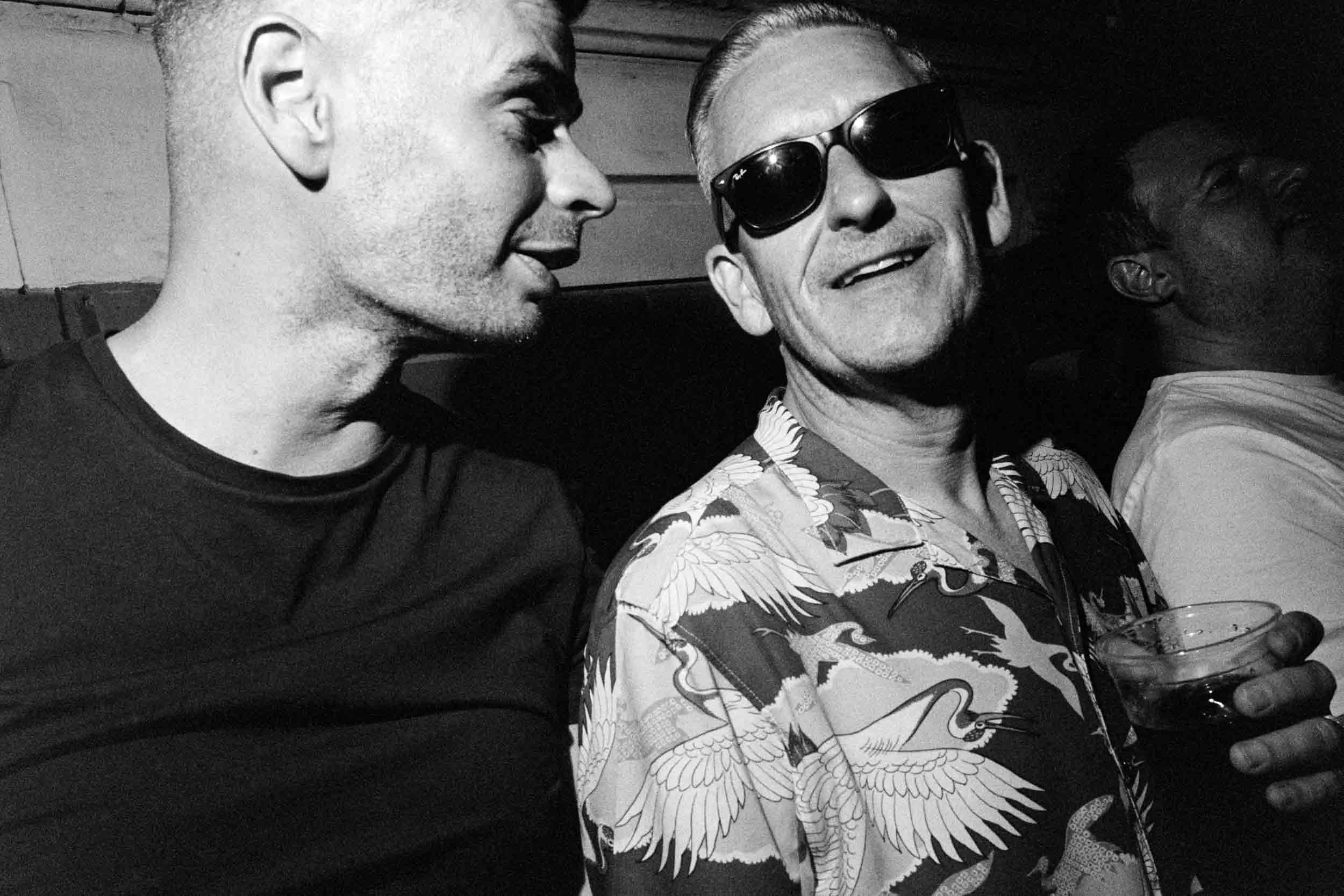 Black and white photo of two men sitting close together, one with sunglasses and a patterned shirt, the other with short hair, both smiling.