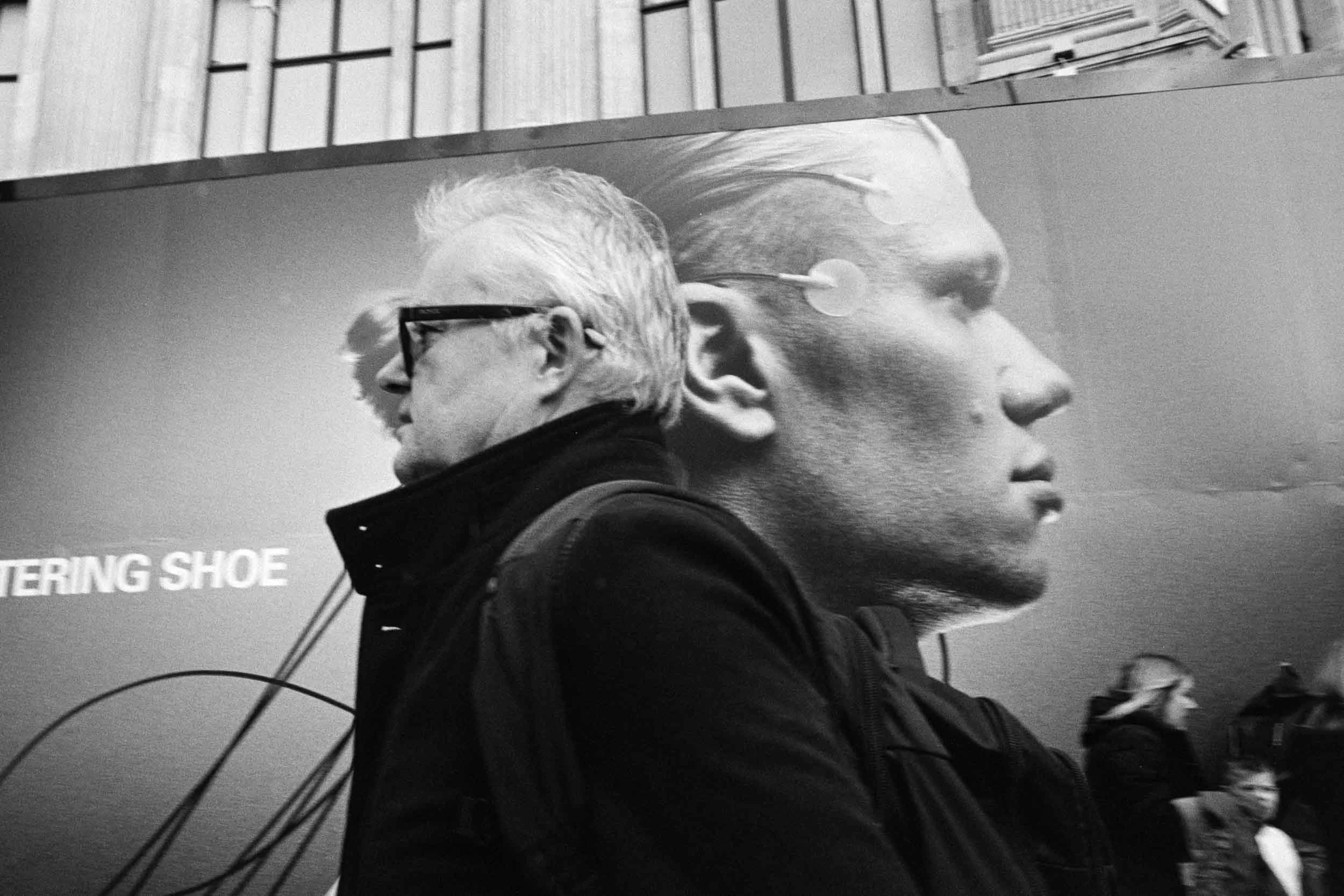 Black and white photograph of two people walking past a large advertisement. The person in the foreground is wearing glasses and a dark jacket, with their face turned to the left. The second person is a large head profile overlapping the first person