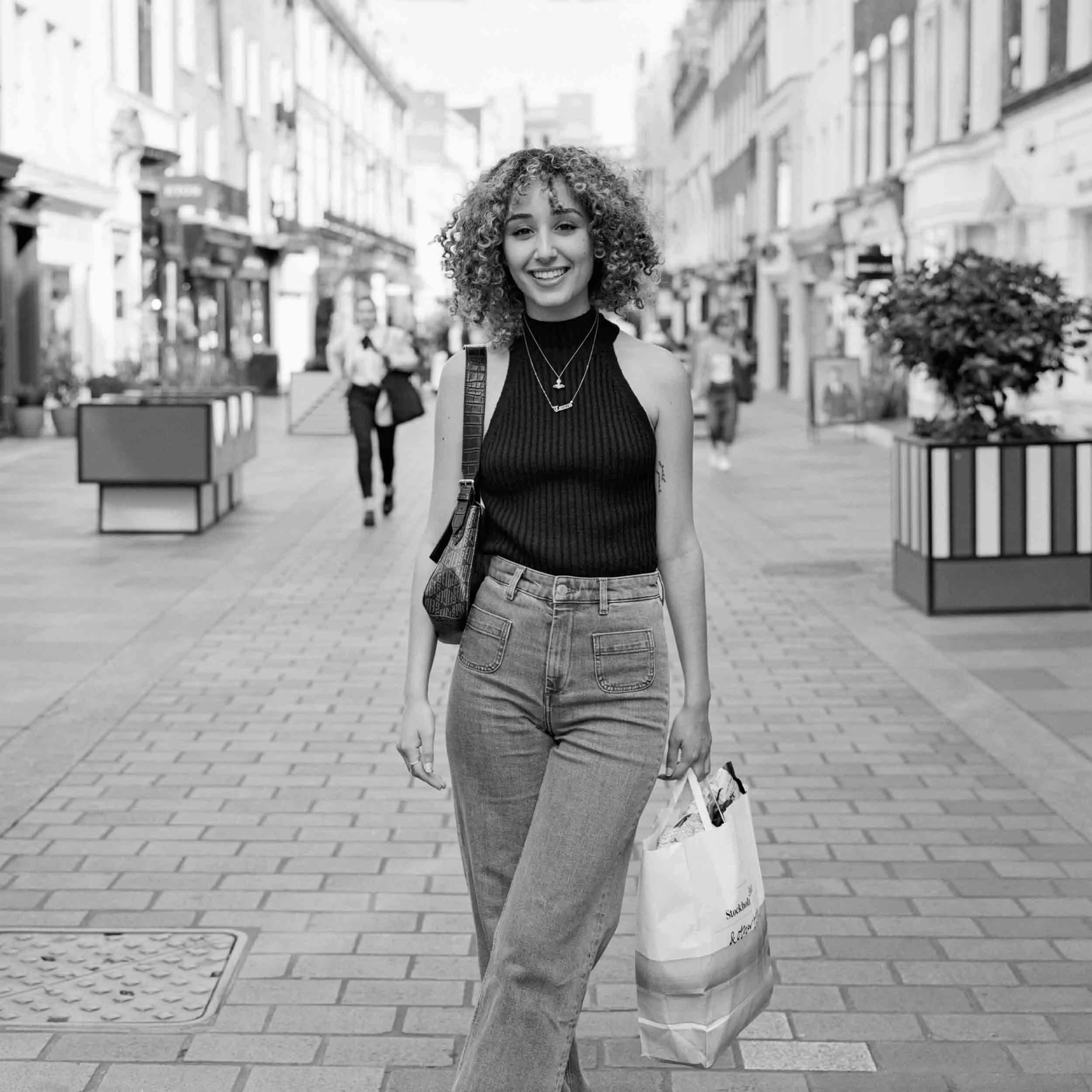 A young woman with curly hair smiling, walking on a city street, carrying a shoulder bag and a shopping bag in a black and white photo.