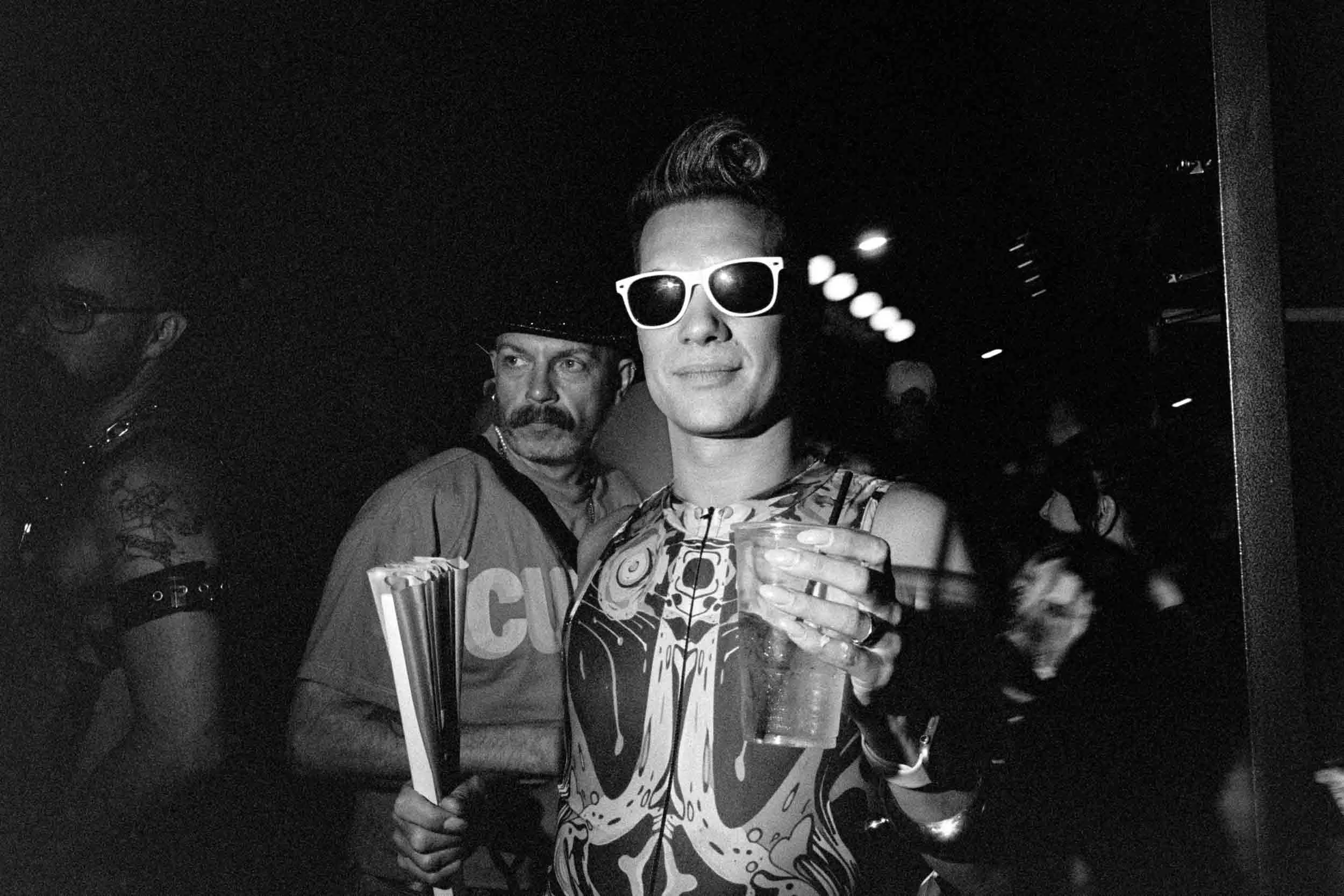 A man wearing sunglasses and a patterned shirt holding a drink, standing with another person wearing a hat, in a dark setting with other people in the background.