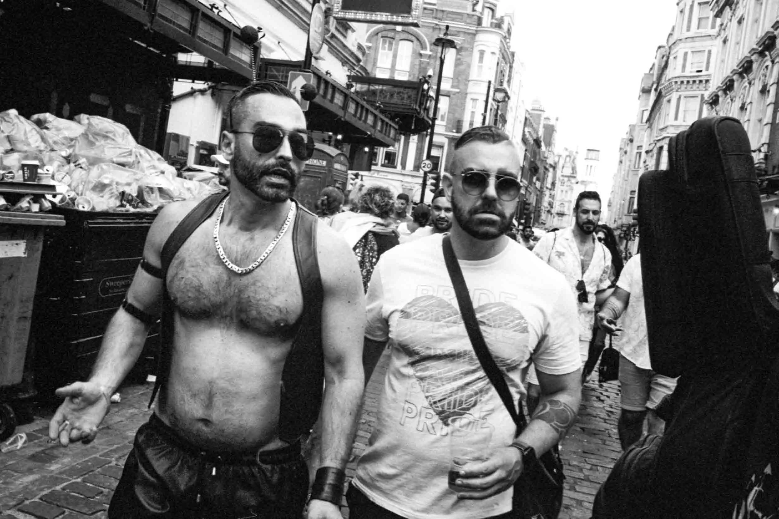 Two men walking down a city street in black and white, with crowds and buildings in the background. One shirtless man with sunglasses and a chain necklace, the other man wearing a short sleeve shirt and sunglasses, both carrying shopping bags.