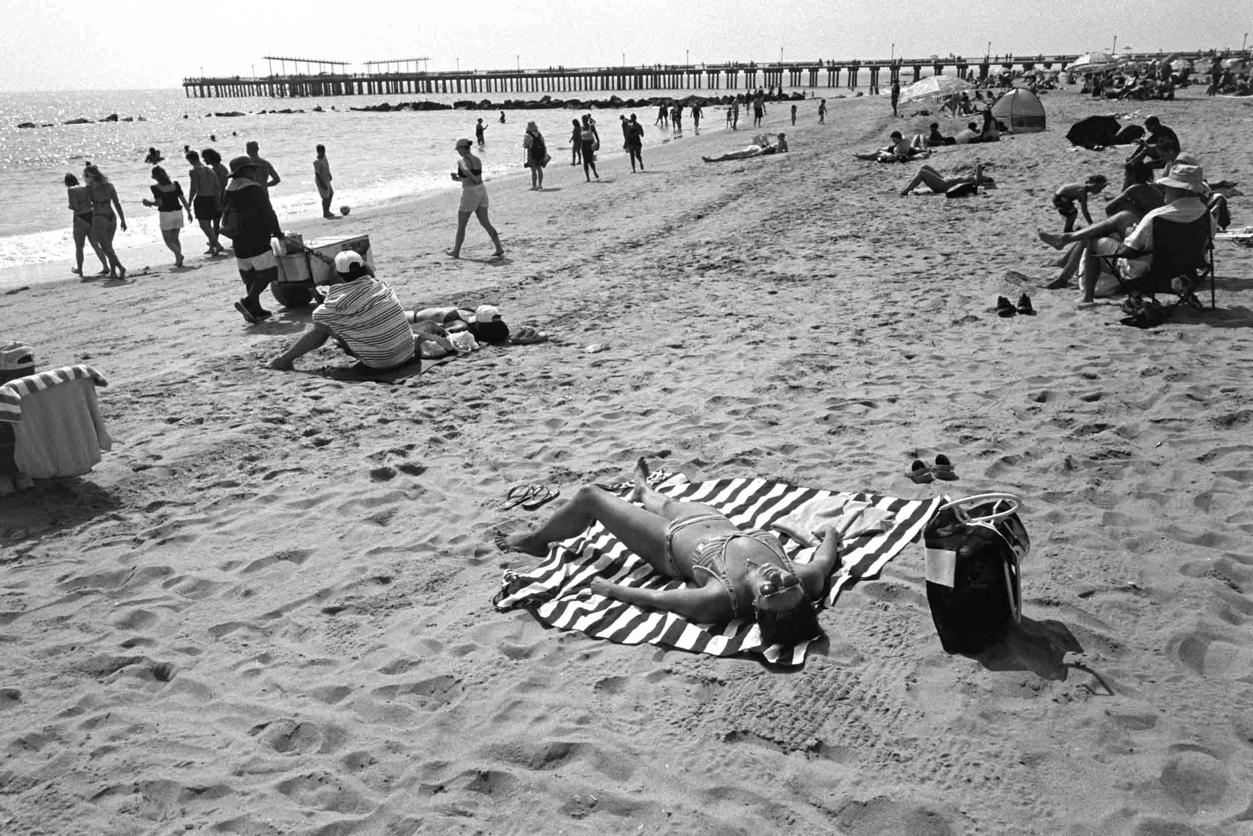 Black and white photo of a crowdedConey Island beach with people relaxing on towels and sitting in chairs, many walking along the shoreline, and a pier extending into the ocean in the background.