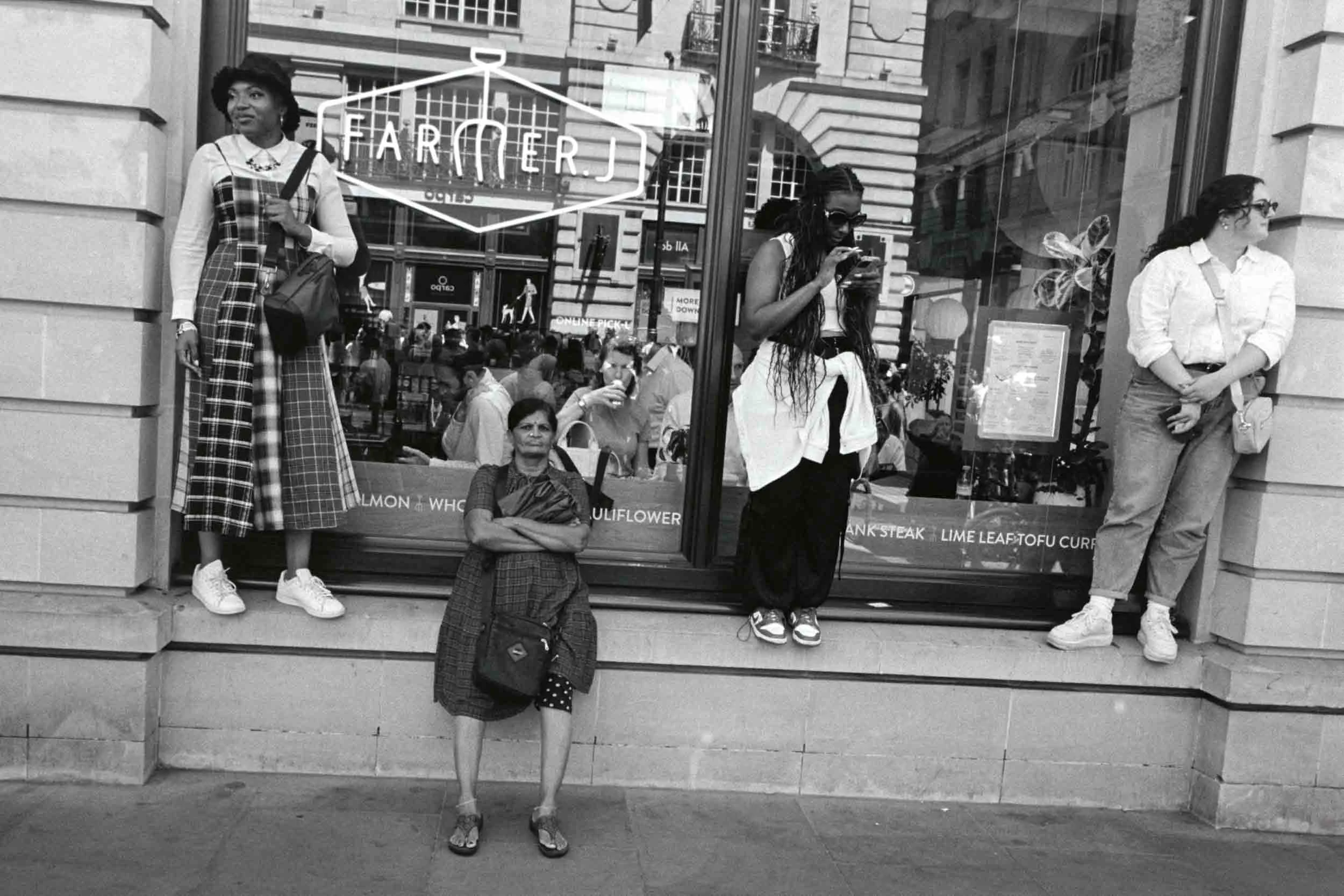 Black and white photo of five women standing and sitting on steps outside a building with large windows. Three women are standing. The woman on the far left wears a plaid dress, white sneakers, and has a purse over her shoulder. The woman on the far 