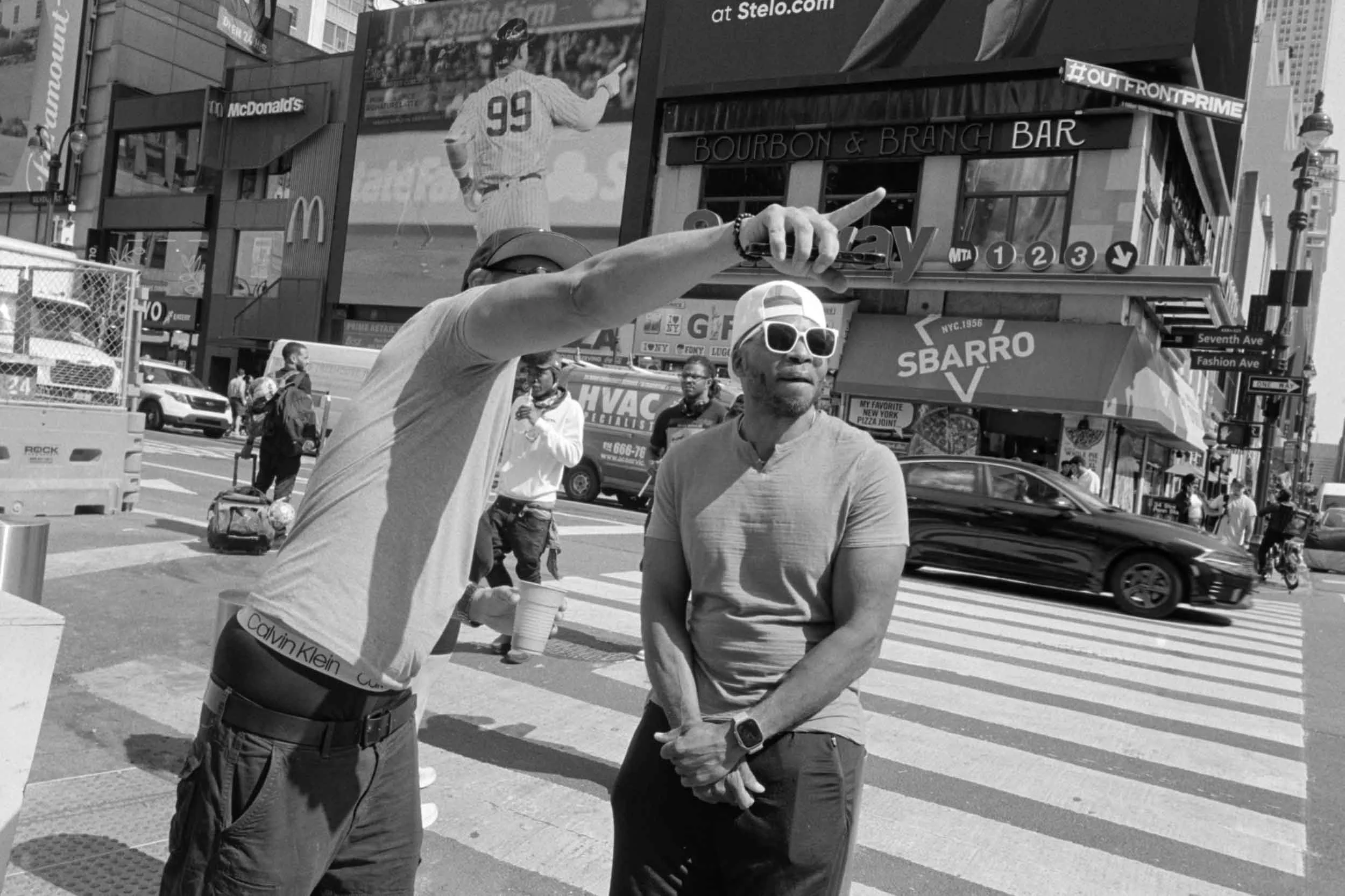 Two men standing on a busy city street in Times Square, New York City, engaged in a conversation. One man is pointing with his finger, and the other man, wearing sunglasses, is looking on. There are advertisements, pedestrians, and moving vehicles ar