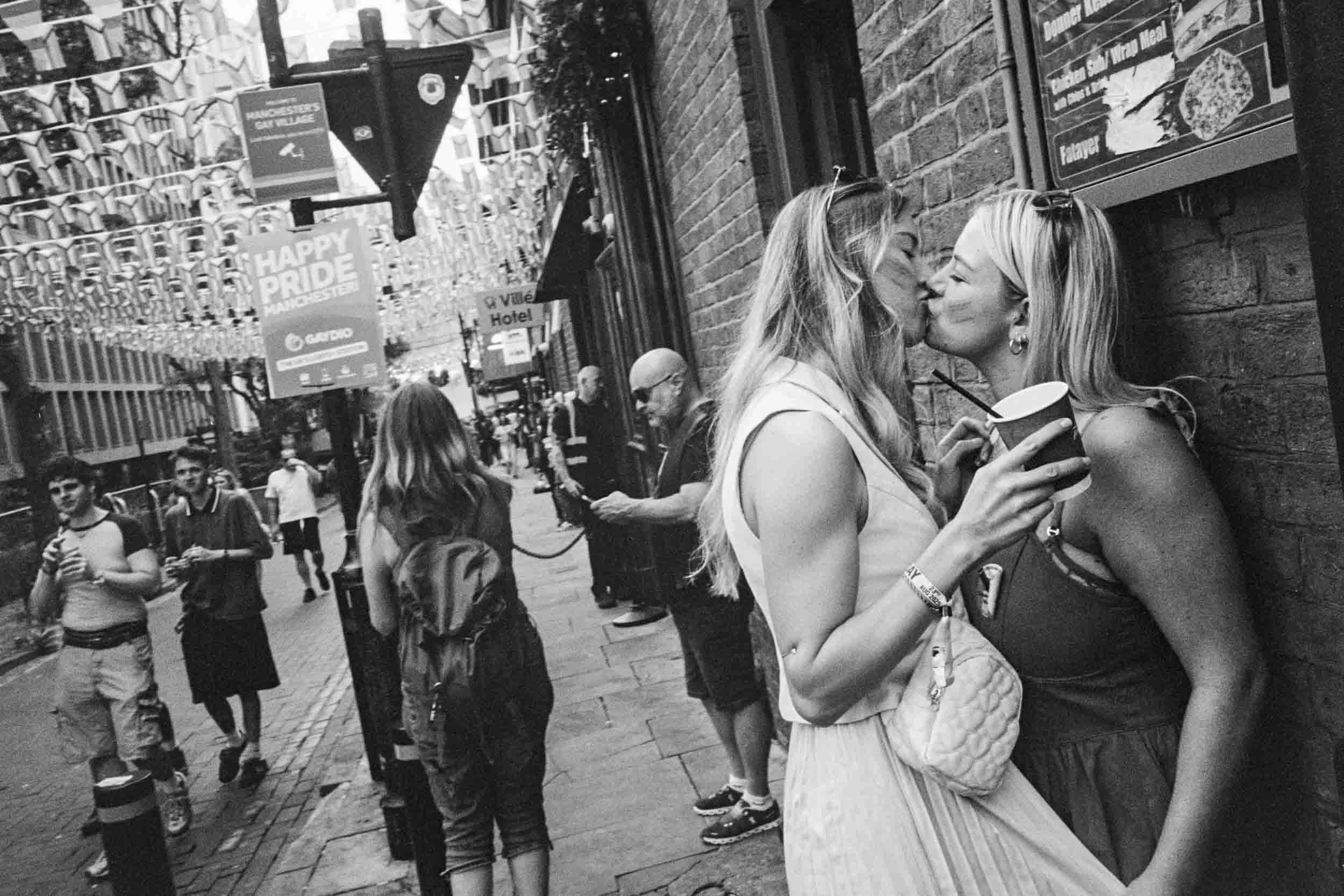 Two women share a kiss on a busy street in Manchester, appearing to celebrate Pride, with various people walking by and colorful banners hanging above.
