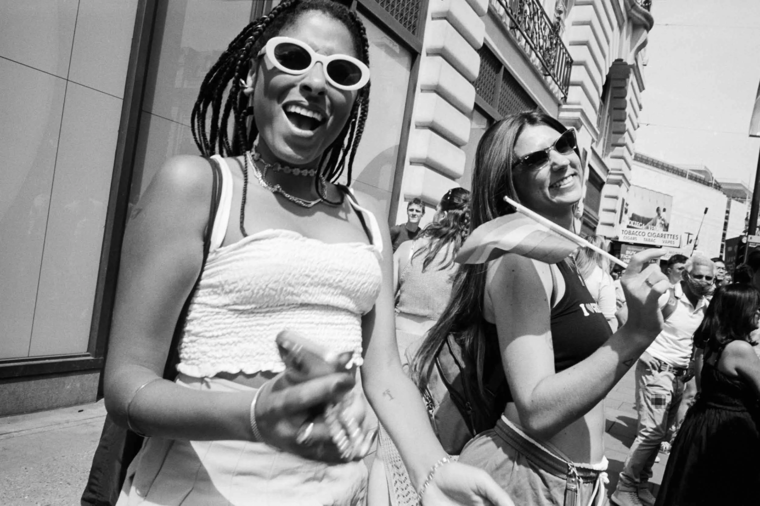 Two women laughing and walking outdoors on a sunny day, wearing sunglasses and casual summer clothing, with a crowd of people and buildings in the background.