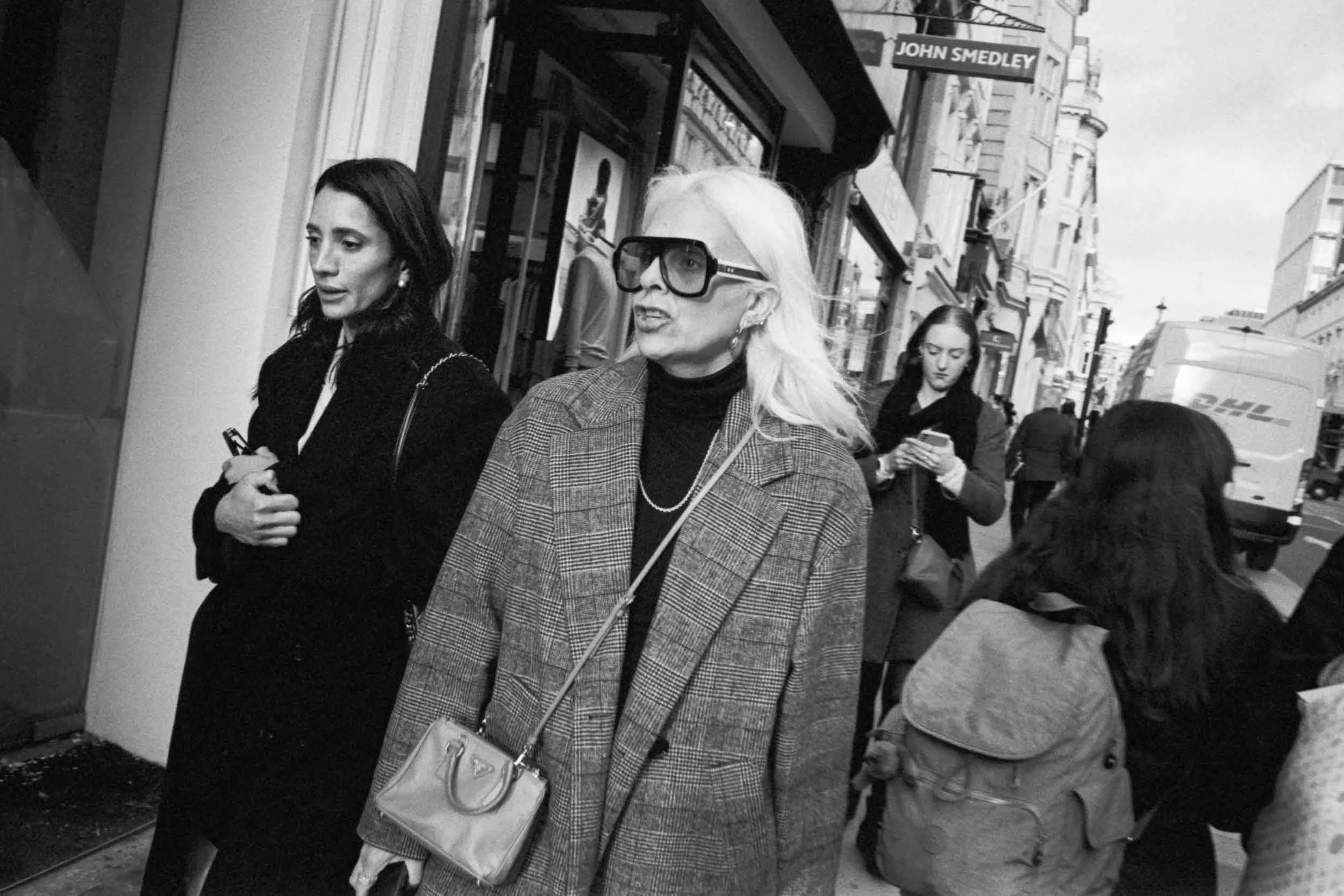 Black and white street scene with three women walking, two women in the foreground, one in the background, buildings with storefronts, a sign reading 'John Smedley' visible above, and a delivery truck on the street.
