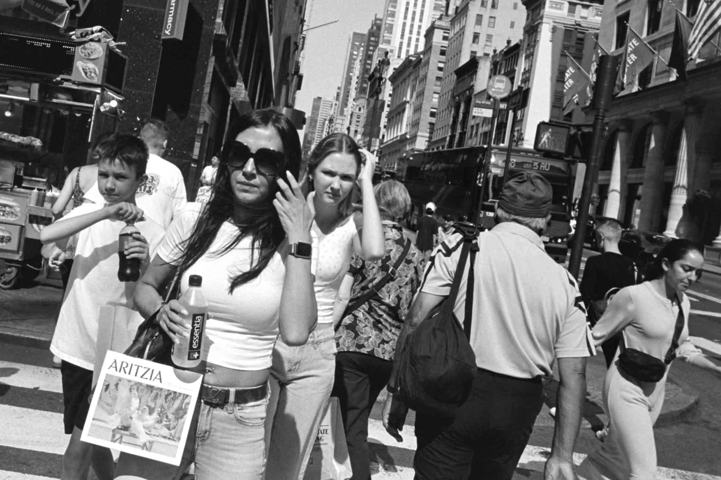 People walking on a busy city street in New York, some carrying shopping bags and wearing casual clothing, with tall buildings, signs, and a trolley in the background.