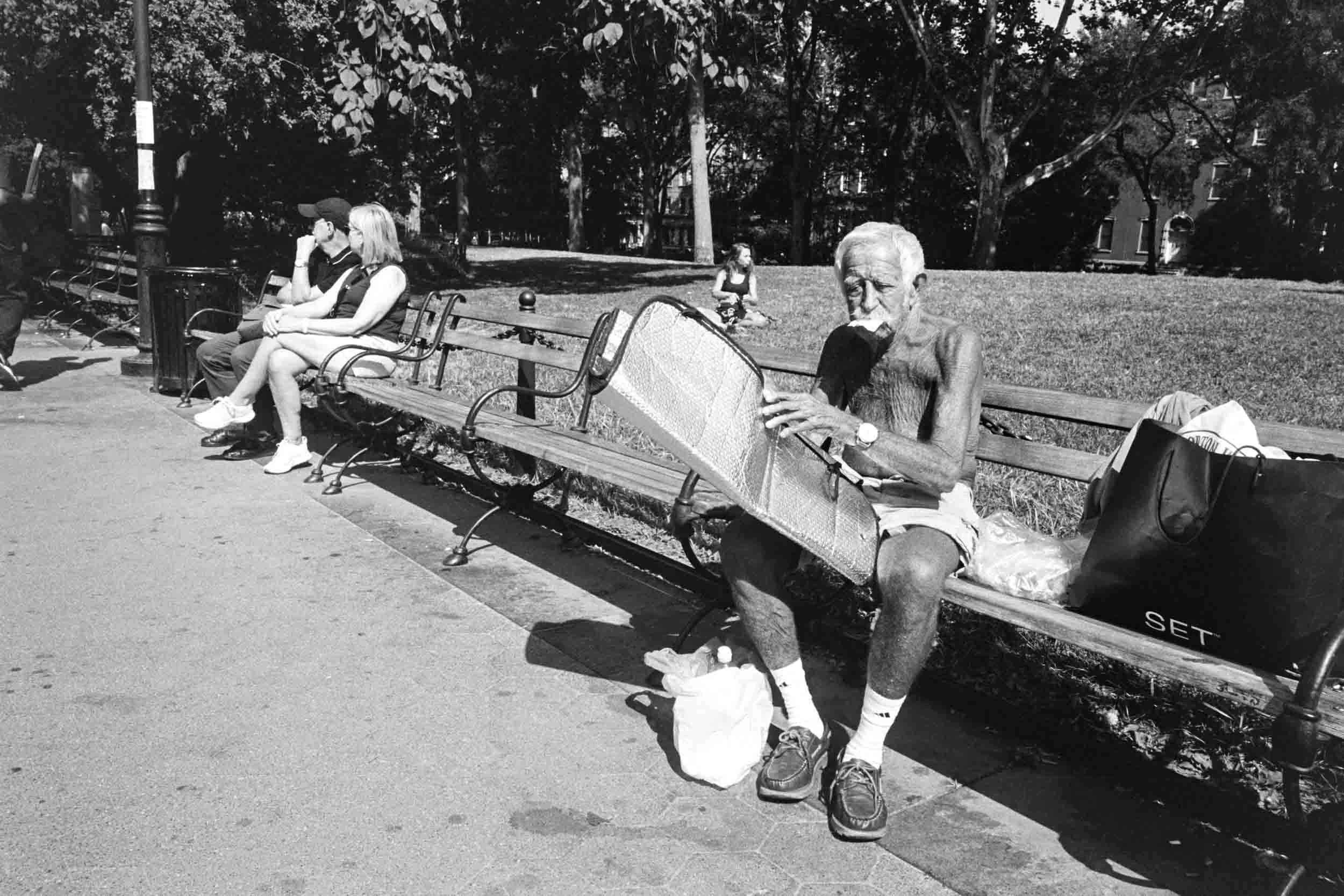 A black and white photo of Washington Square Park with several people sitting on benches, including an elderly man reading a newspaper. There are trees and buildings in the background.