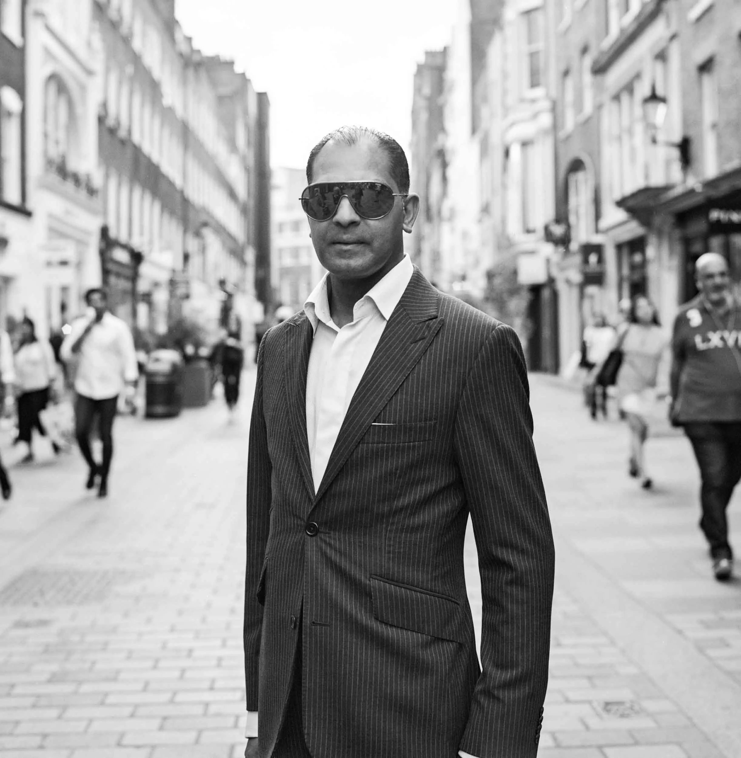A man in a pinstripe suit and sunglasses walking down a city street with buildings on either side.