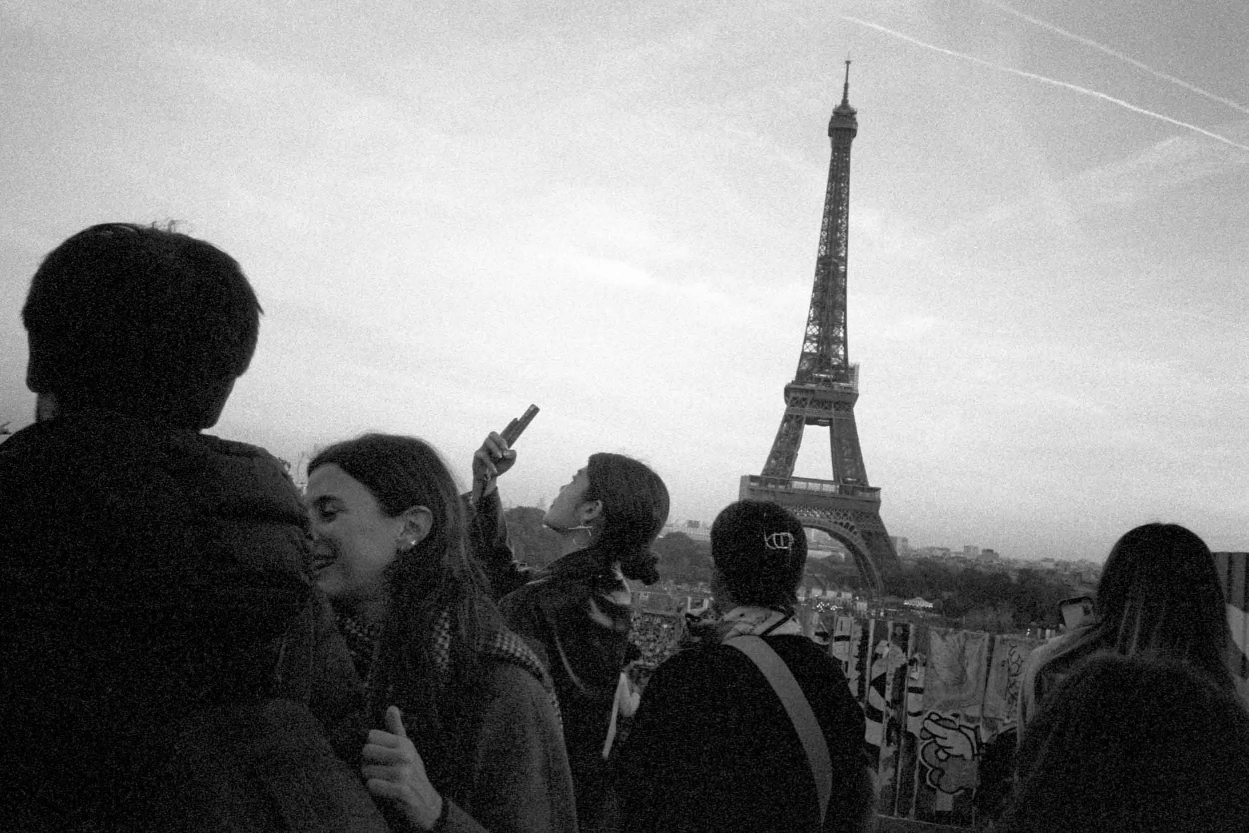 A black and white photo of a crowd of people with the Eiffel Tower in the background, some people are holding up smartphones, and a few are smiling.