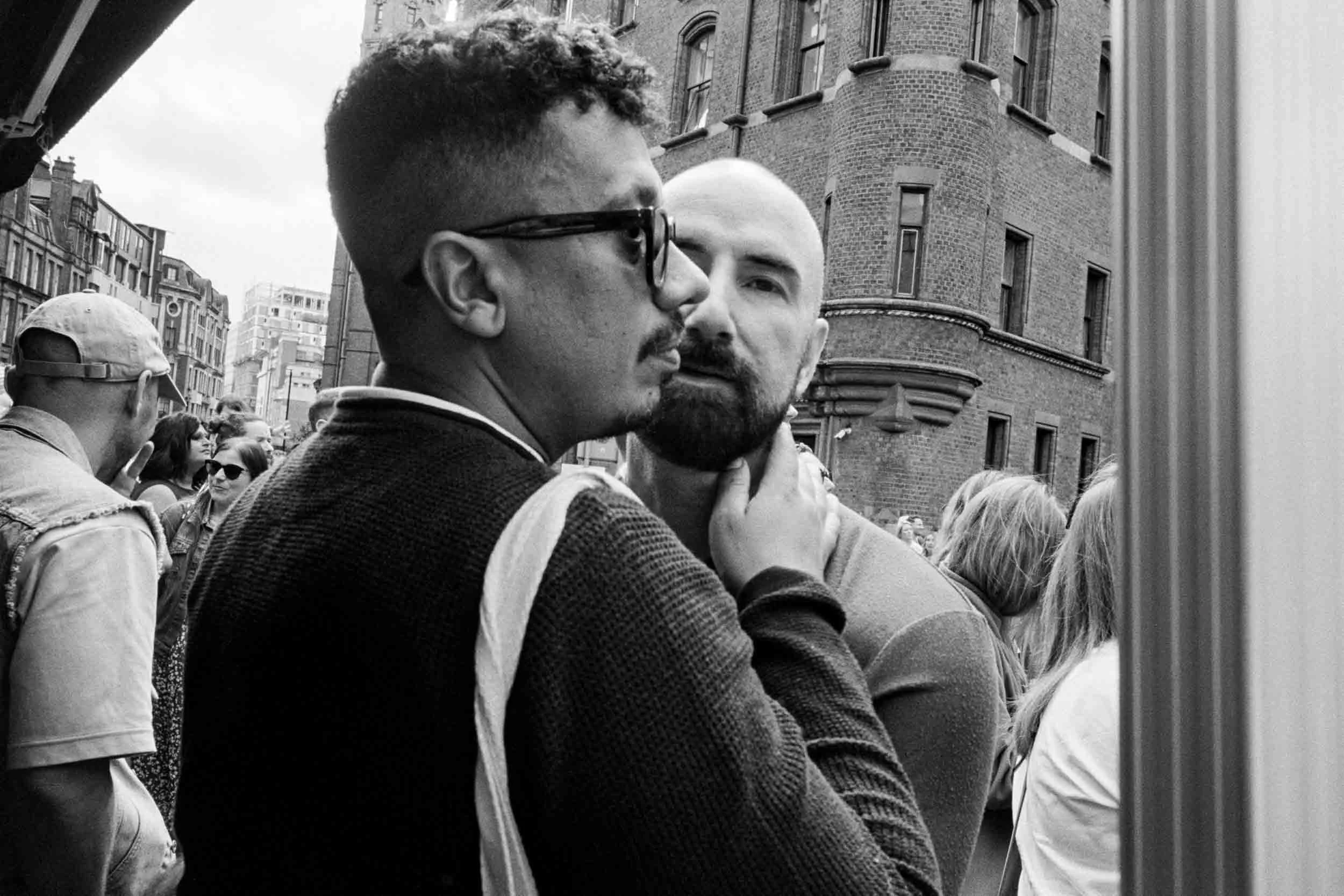 Two men face each other closely in an urban street scene with city buildings in the background; one man is wearing glasses and a jacket, and the other has a shaved head and beard.