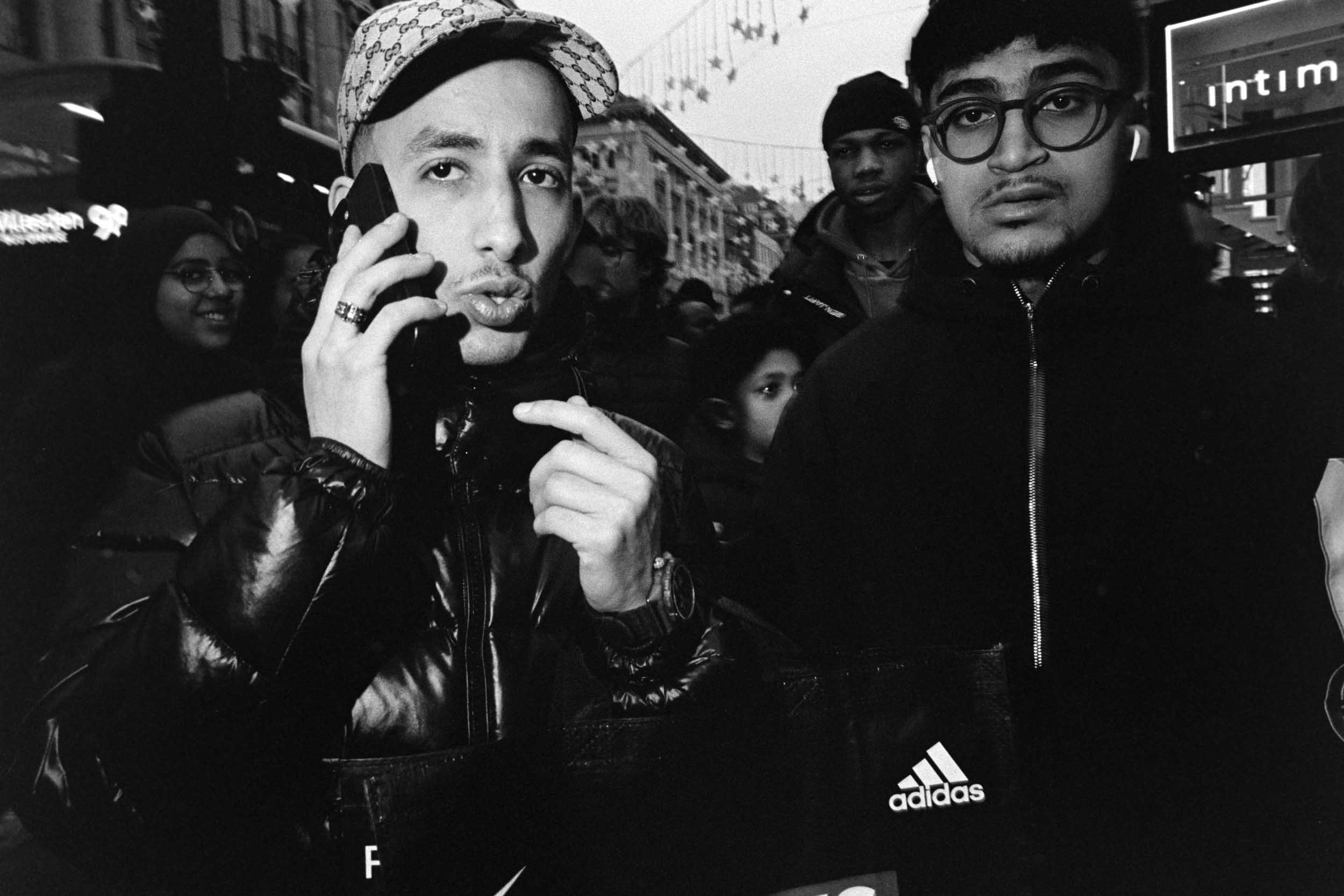 Black and white photo of three young men in outdoor urban setting. The man on the left has light skin, a hat with a Gucci pattern, and is talking on a phone. The man on the right has darker skin, glasses, and a hoodie, looking towards the camera. The