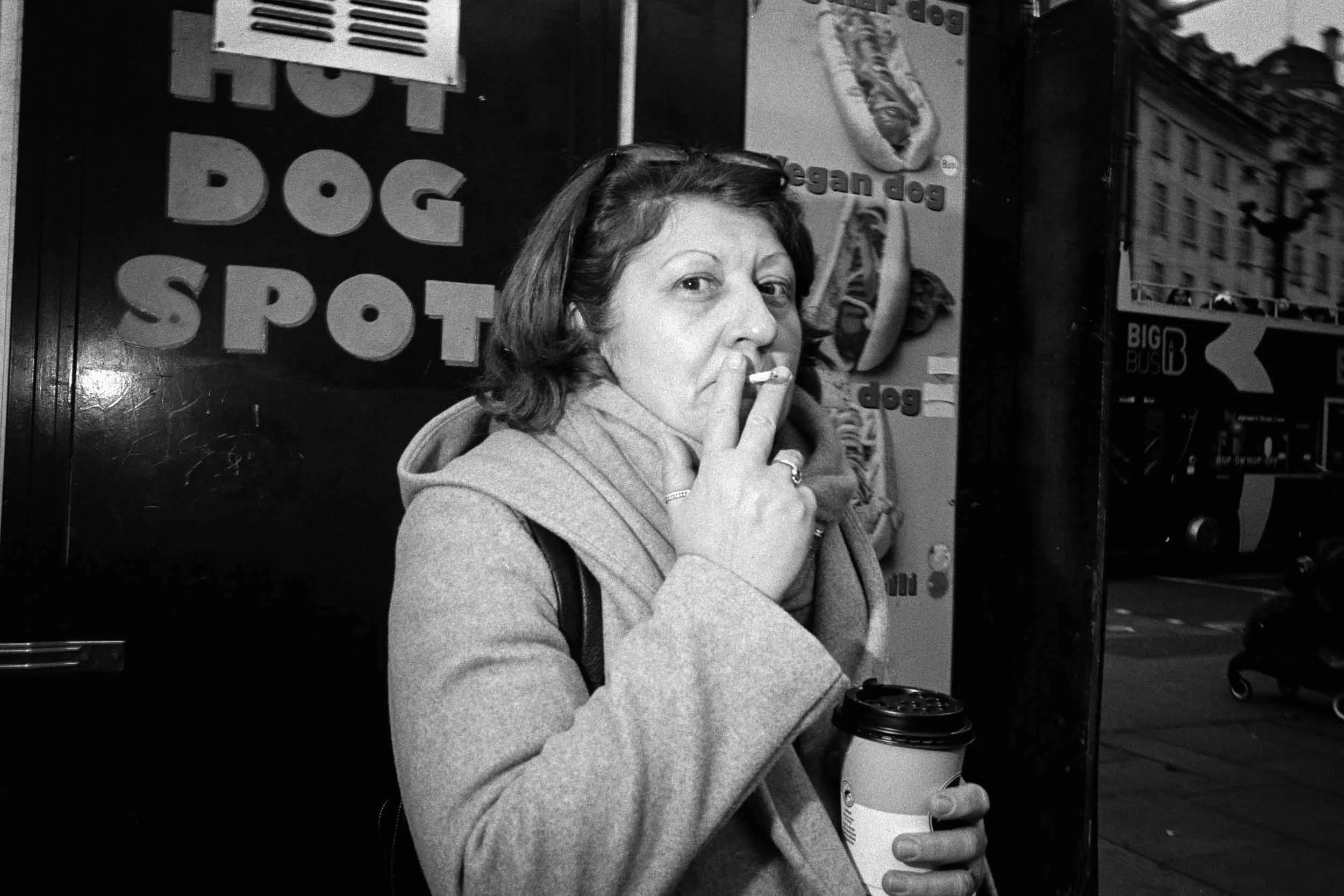 A woman with shoulder-length hair and glasses on her head, wearing a coat, is sitting outdoors, smoking a cigarette and holding a to-go coffee cup. She appears to be in an urban setting with some signs and buildings in the background.