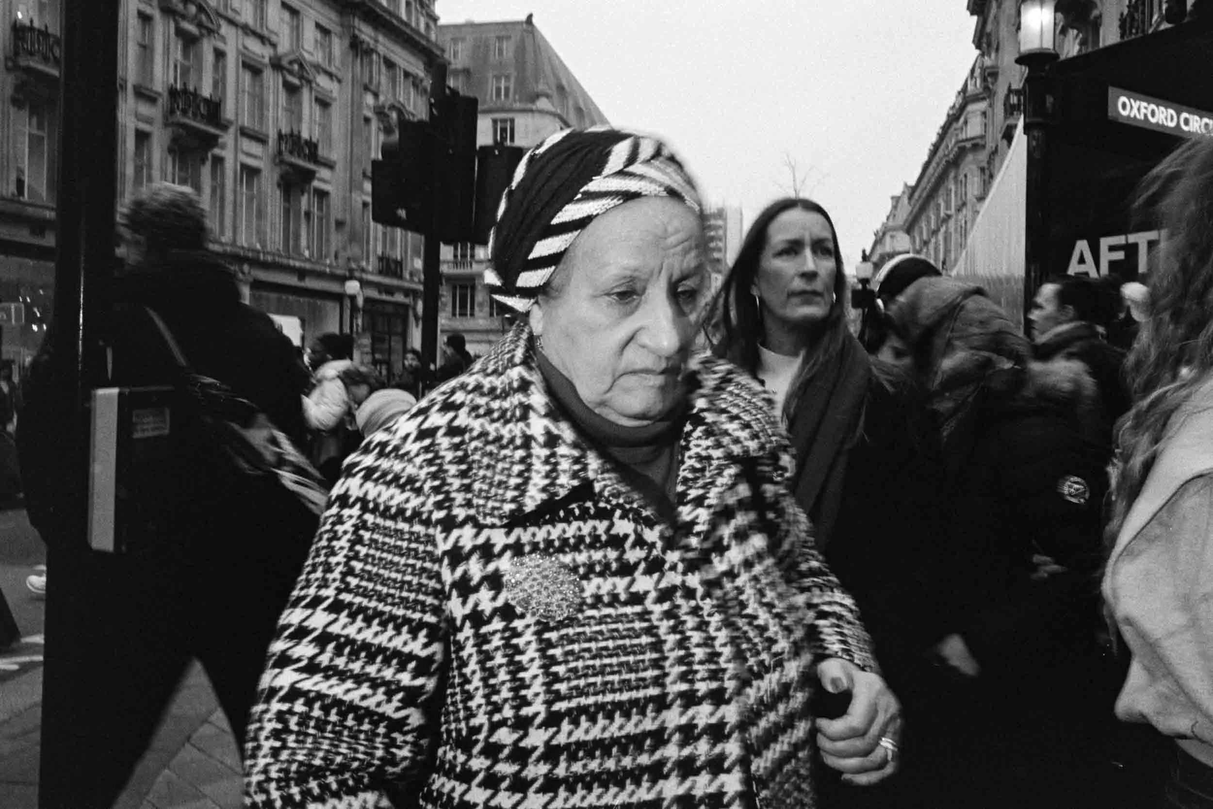 Black and white photo of an older woman wearing a patterned coat and a headscarf, walking in a busy city street with pedestrians and historic buildings, in front of a sign that reads 'Oxford Circus'.