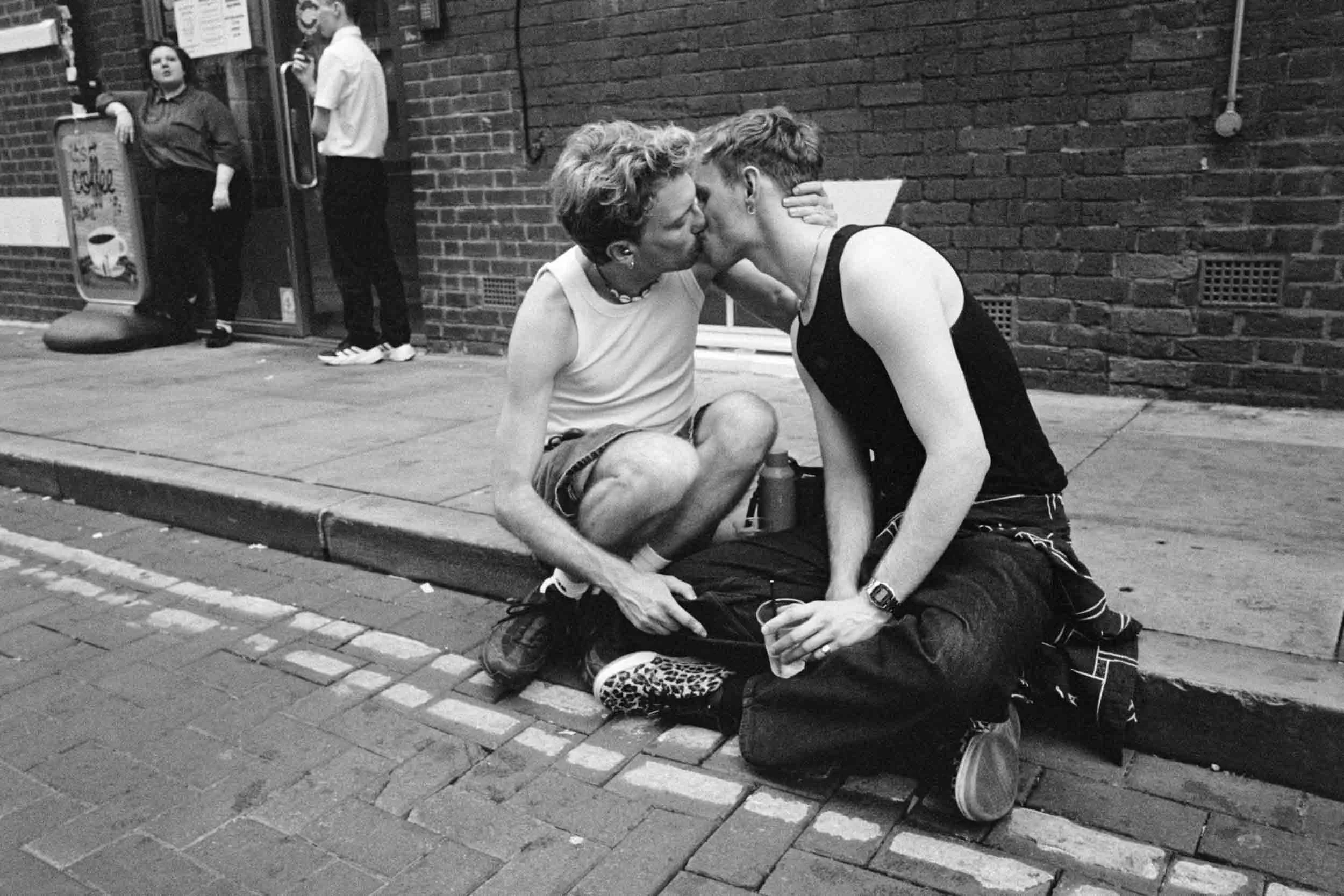 Two people are sitting on the sidewalk, sharing a kiss, with one holding a drink. There are two other individuals standing near a brick wall in the background, one leaning on a doorframe and the other smoking. The scene is at Manchester Pride Canal S