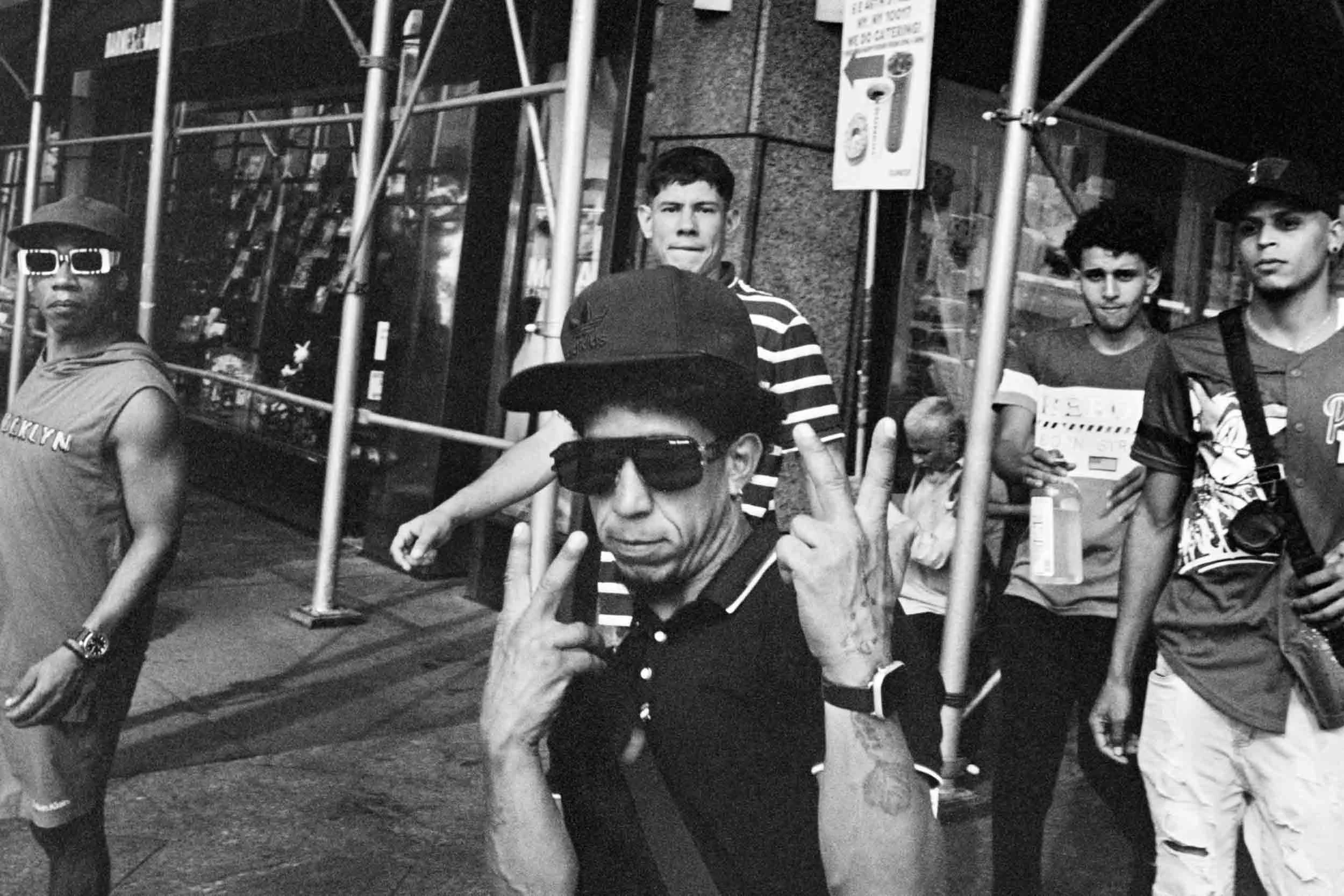A group of young men standing behind a metal scaffold on a city sidewalk. One man is making a peace sign with both hands, and another is holding a drink; the others are standing or walking. The scene is in black and white.