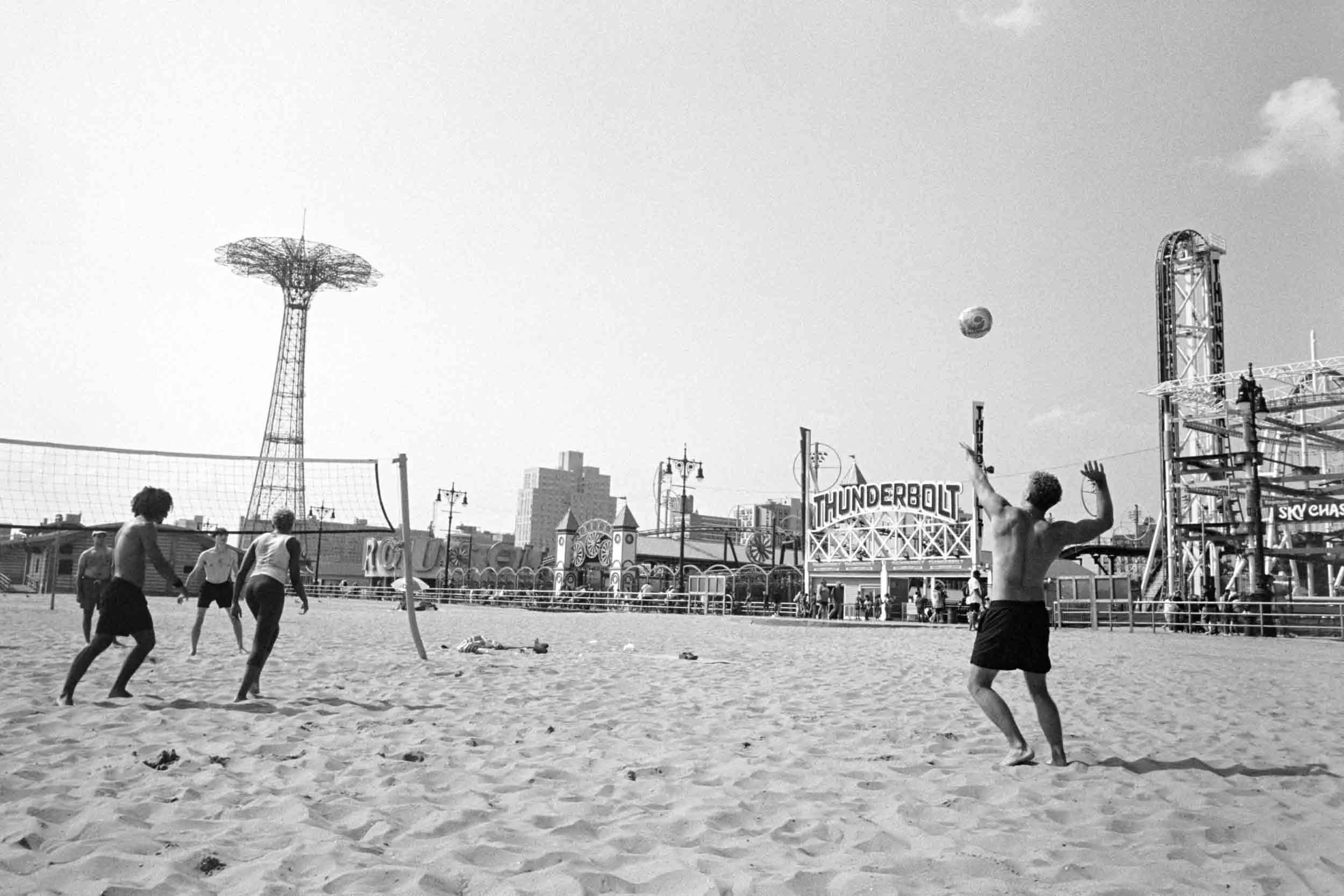People playing volleyball on the beach at Coney Island with amusement park rides and structures in the background, including a tall tower and a sign that says 'THUNDERBOLT'.