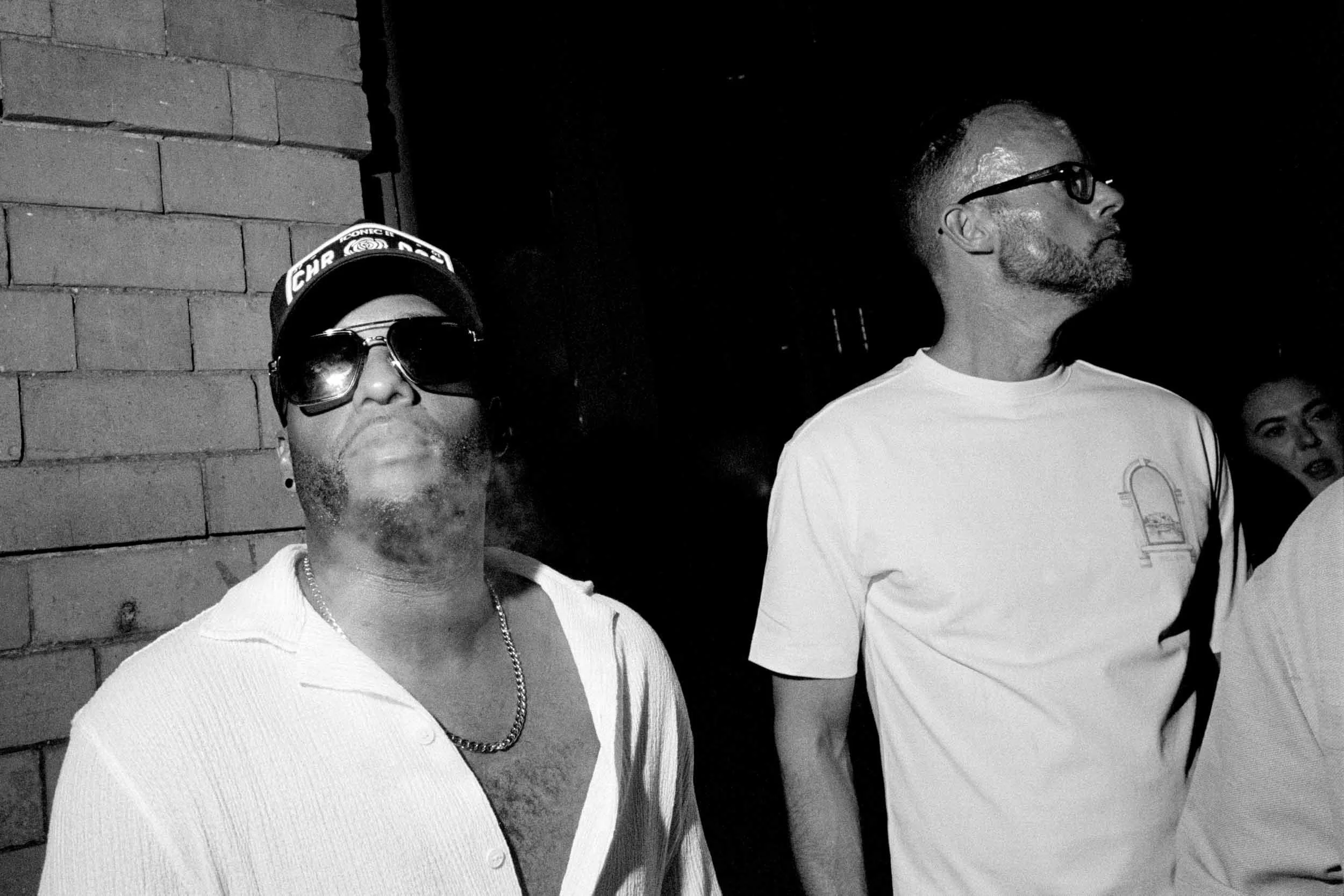 Two men stand side by side against a brick wall at night during Manchester Pride. The man on the left wears sunglasses, a hat with text, and a chain necklace. The man on the right has glasses, a beard, and a white t-shirt.