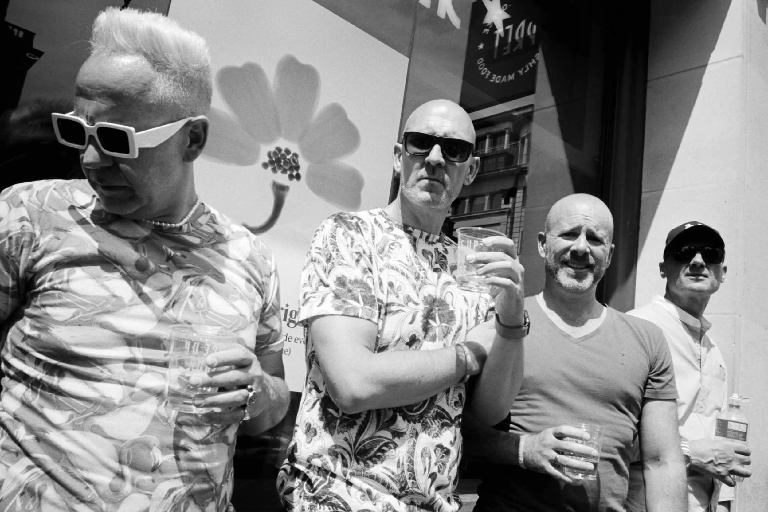A group of men standing outdoors at London Pride, some wearing sunglasses and holding drinks, with a large flower mural and buildings in the background.