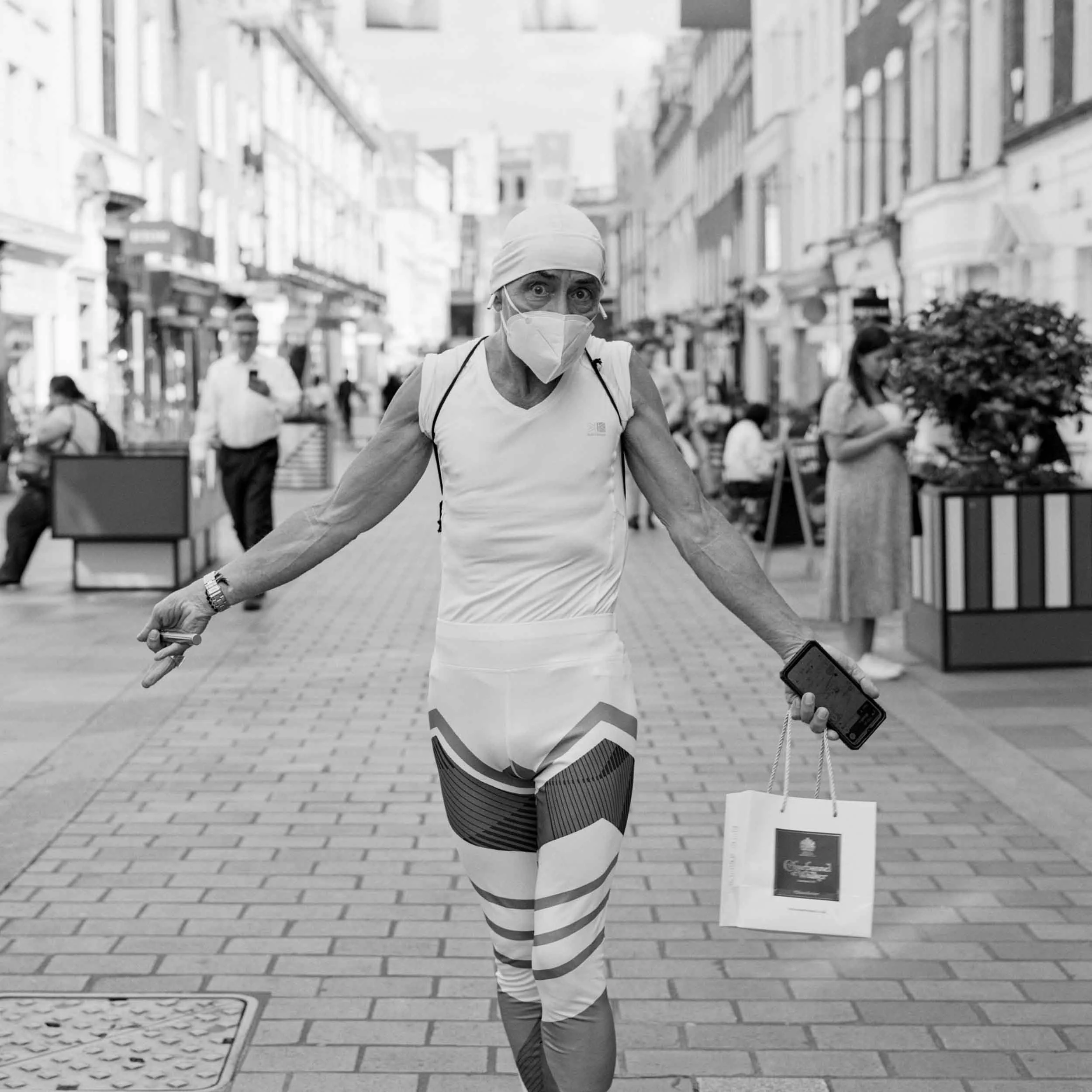 A woman in athletic clothing and a face mask walking outdoors on a city street with shops and other pedestrians, holding a smartphone and a walking stick.