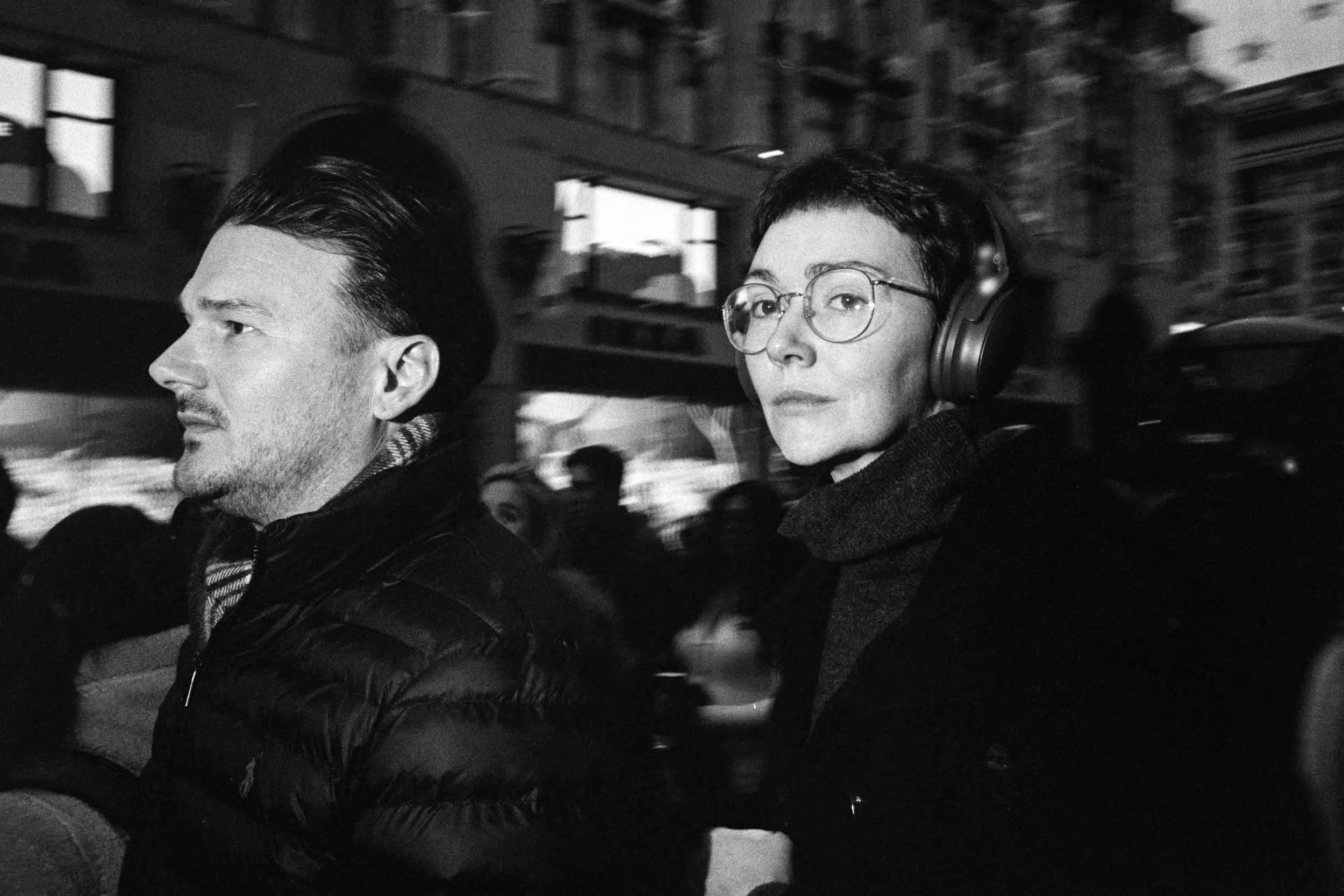 Black and white photo of a man and a woman riding in a moving vehicle at night. The man has dark, slicked-back hair and a mustache,while the woman has short dark hair, glasses, and headphones, looking to the right.