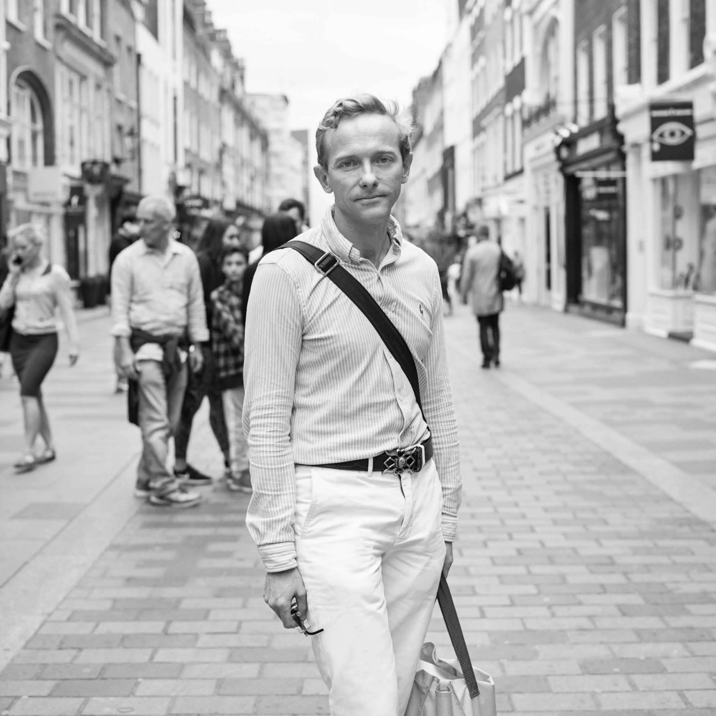 A black and white photo of a man walking on a busy city street with buildings and storefronts, carrying a shoulder bag and holding a cane.