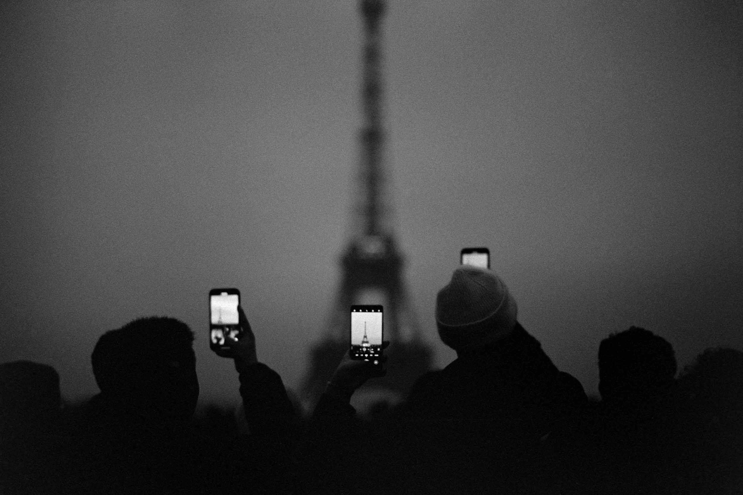 A black and white photo of a crowd holding up phones to take pictures or videos of a the Eiffel Tower, Paris with one phone showing the tower's silhouette against the sky.