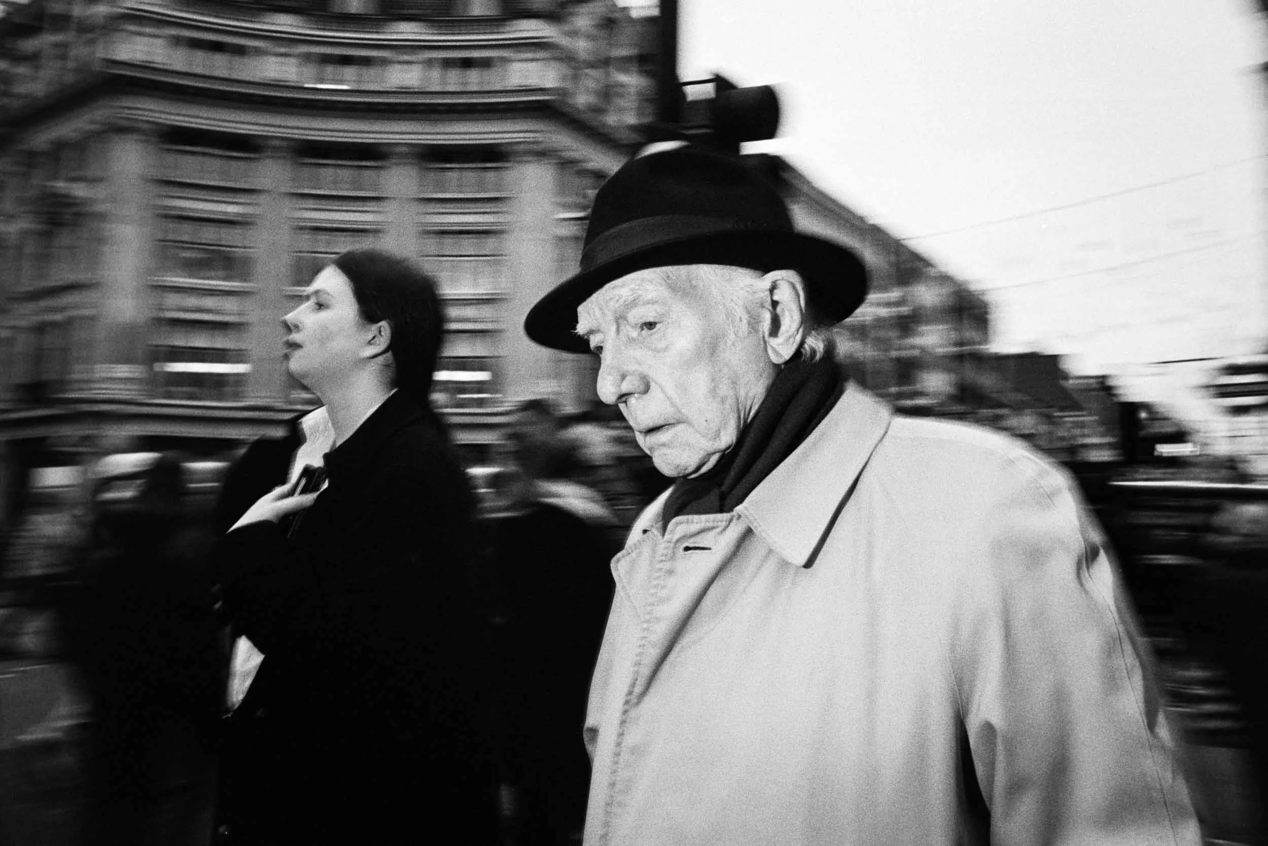 Black and white photo of an elderly man and a woman walking in Oxford Circus London with blurred buildings and traffic in the background, both in profile, the man wearing a hat and coat, the woman with dark hair.