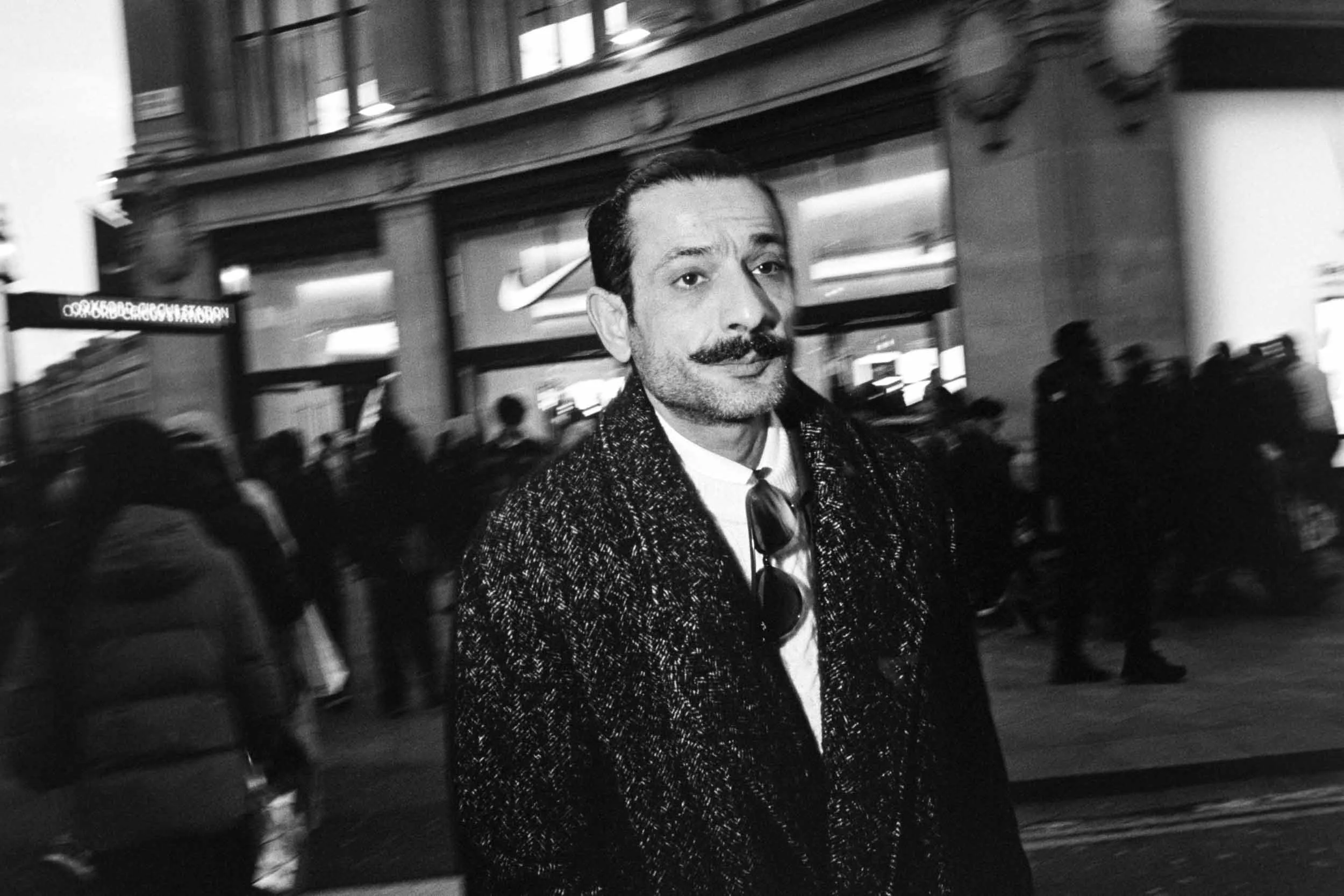 Black and white photo of a man with dark hair, a mustache, and a beard, wearing a coat and tie, standing on a busy city street.