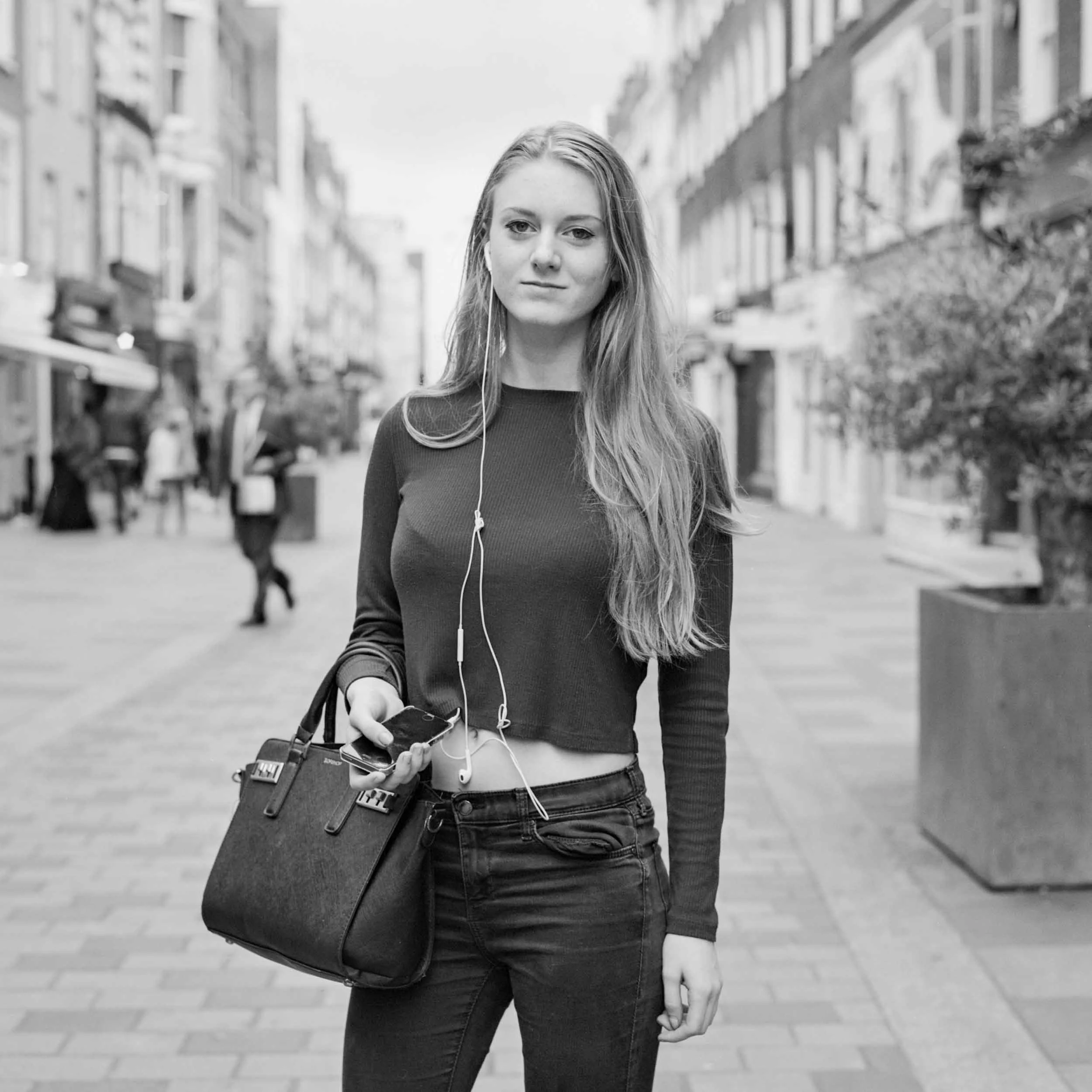 A young woman with long hair wearing a long sleeve top and jeans, holding a skateboard in one hand and an iPhone with earphones plugged in while walking on a city street.