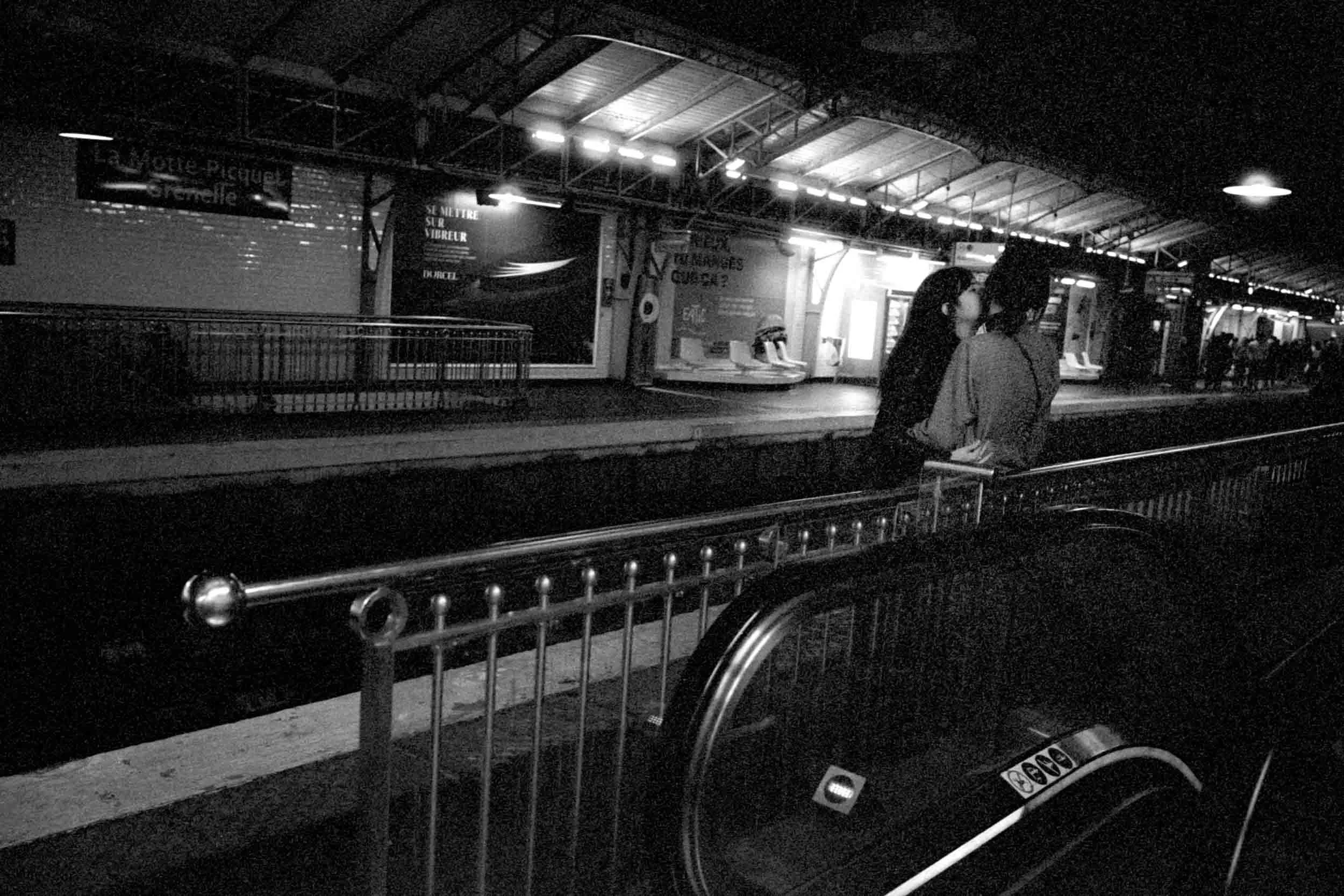 Nighttime scene at an outdoor station with two people standing near a luggage trolley, illuminated advertisements on the wall, and other people waiting in the background.