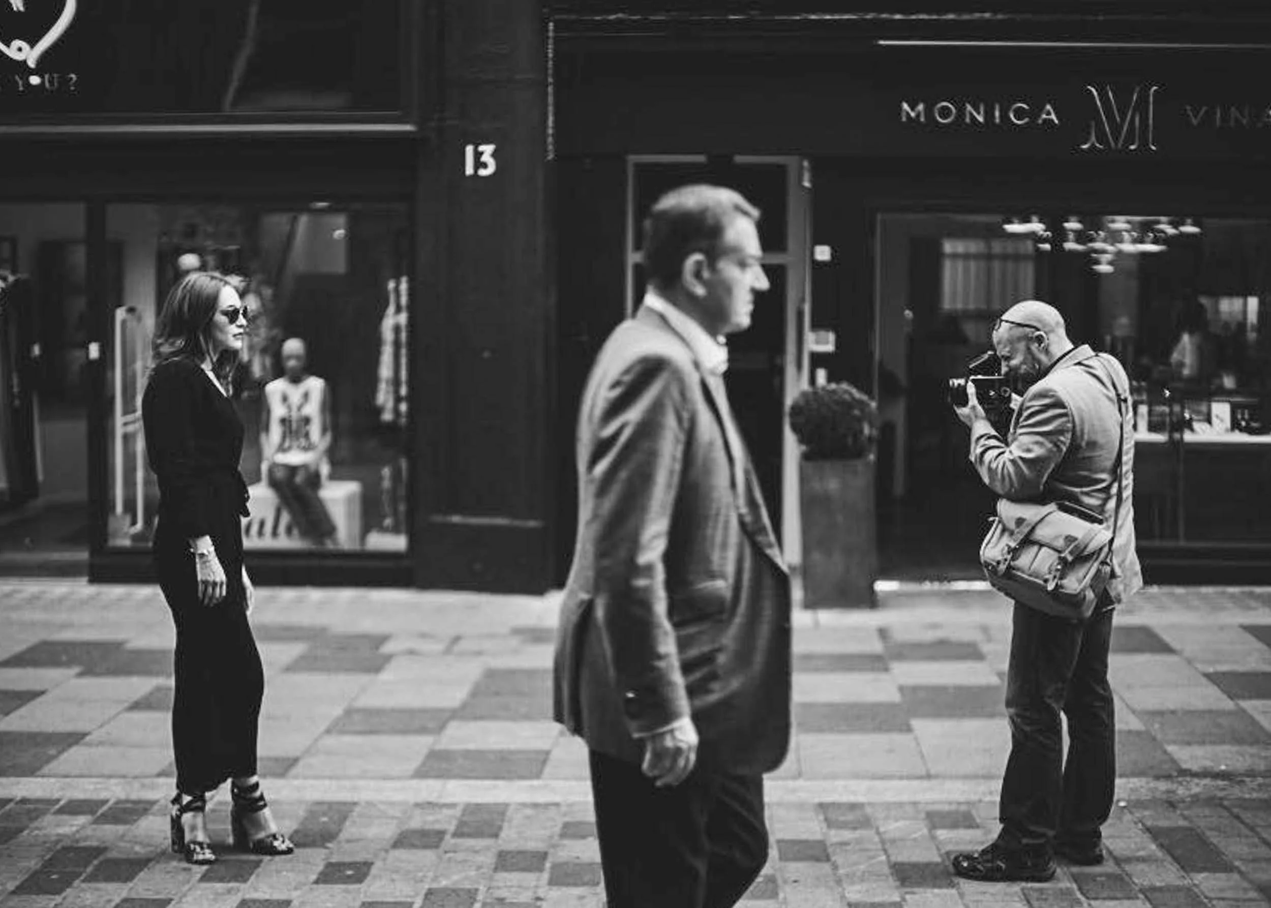 Black and white photo of four people crossing a city street in front of a store named Monica. Two men are walking in the foreground, one man is looking down, and a woman with sunglasses is standing on the sidewalk.