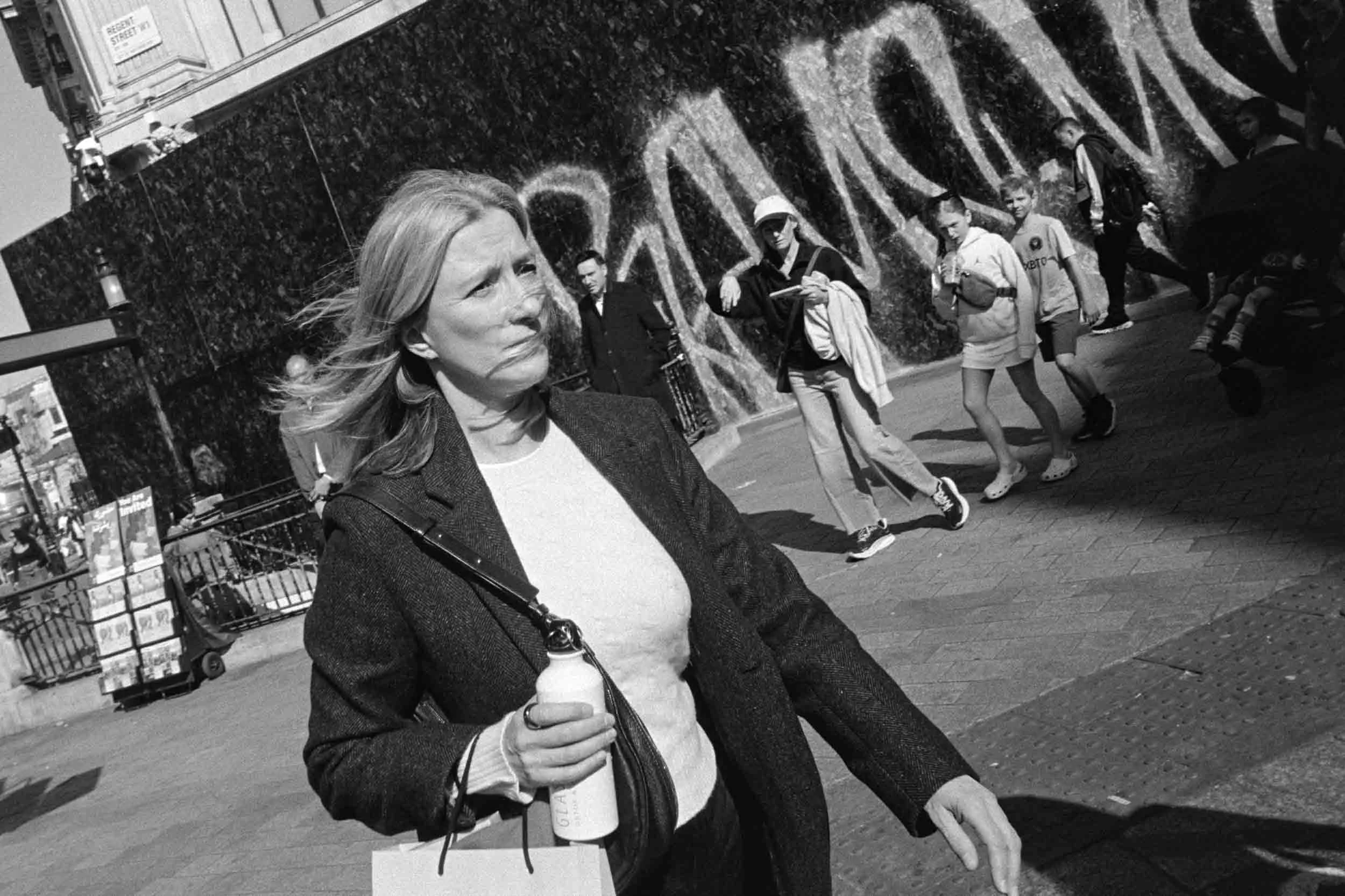 Black and white photo of a woman with long hair wearing a blazer, holding a coffee cup, standing in front of a graffiti-covered wall, with several people in the background walking and standing in an urban street.