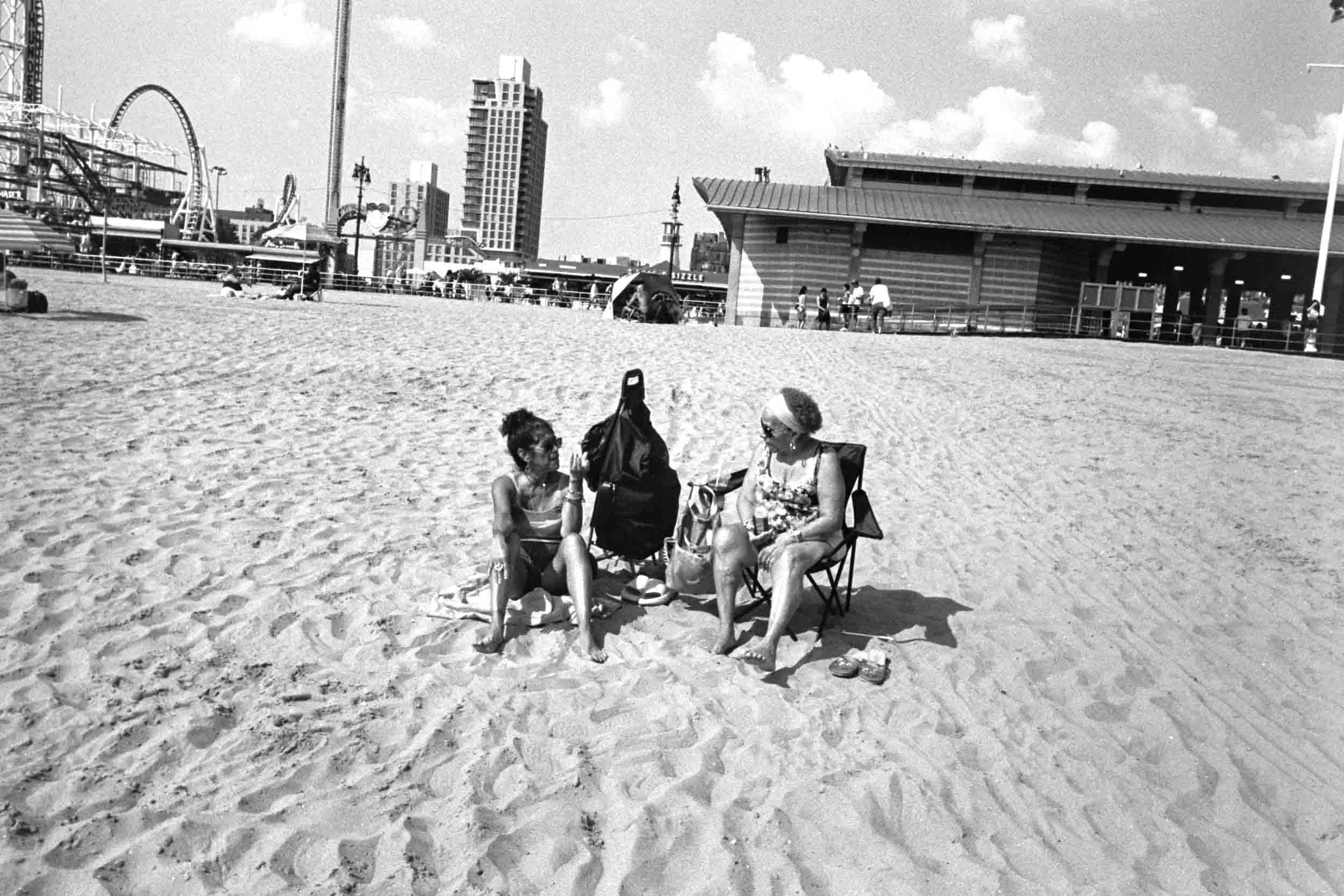 Two women sitting on beach chairs and talking at the beach in Coney Island, with a boardwalk, amusement park rides, and buildings in the background in a black and white photo.
