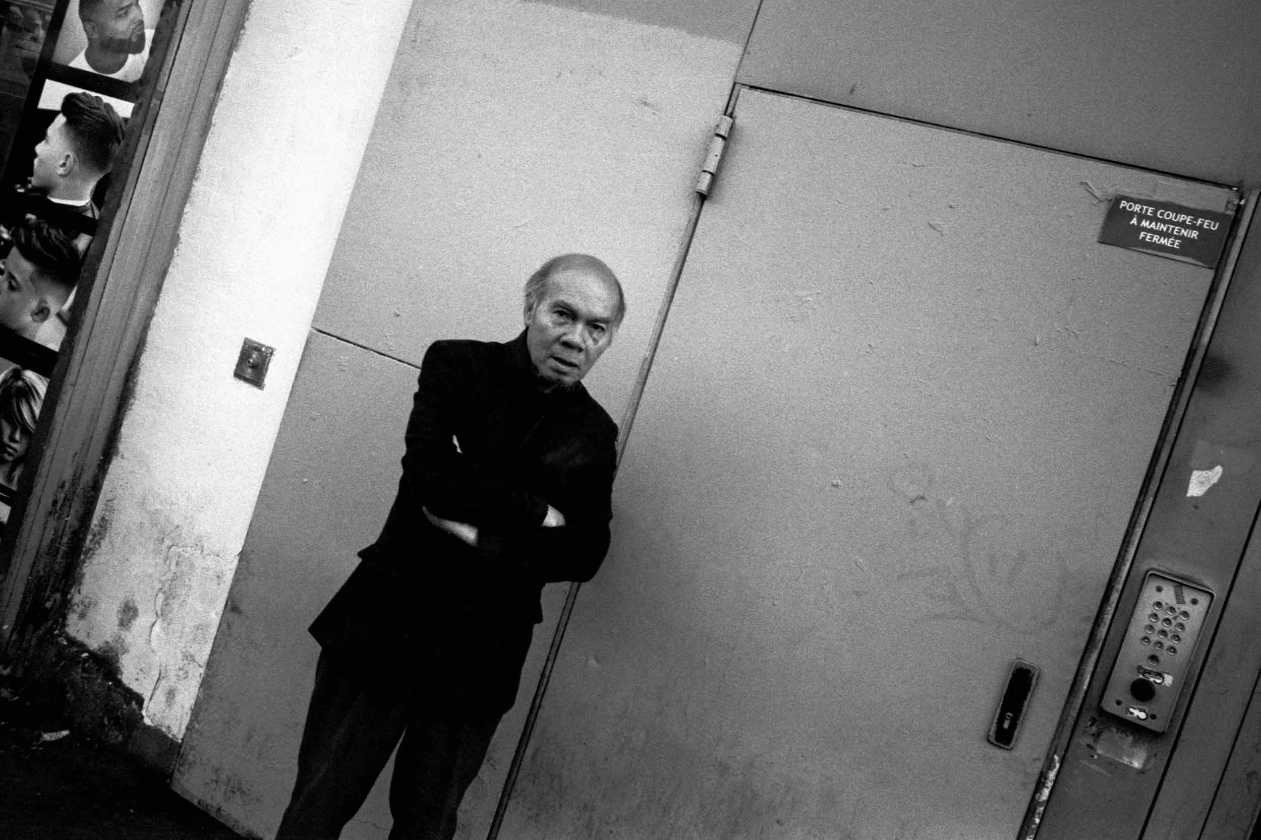 A black and white photo of a man standing with his arms crossed, looking at the camera, in front of a plain wall with a closed door. There is a sign on the door that indicates it is a room that must be kept closed, written in both French and English.