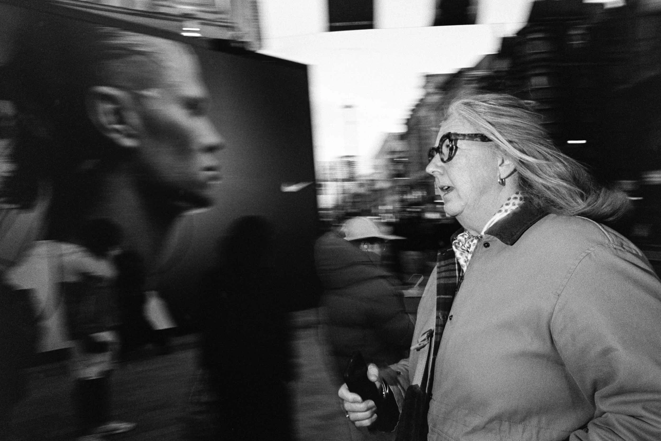 A woman with glasses and long hair walking on a busy street, looking to the right, while several blurred people move past her.