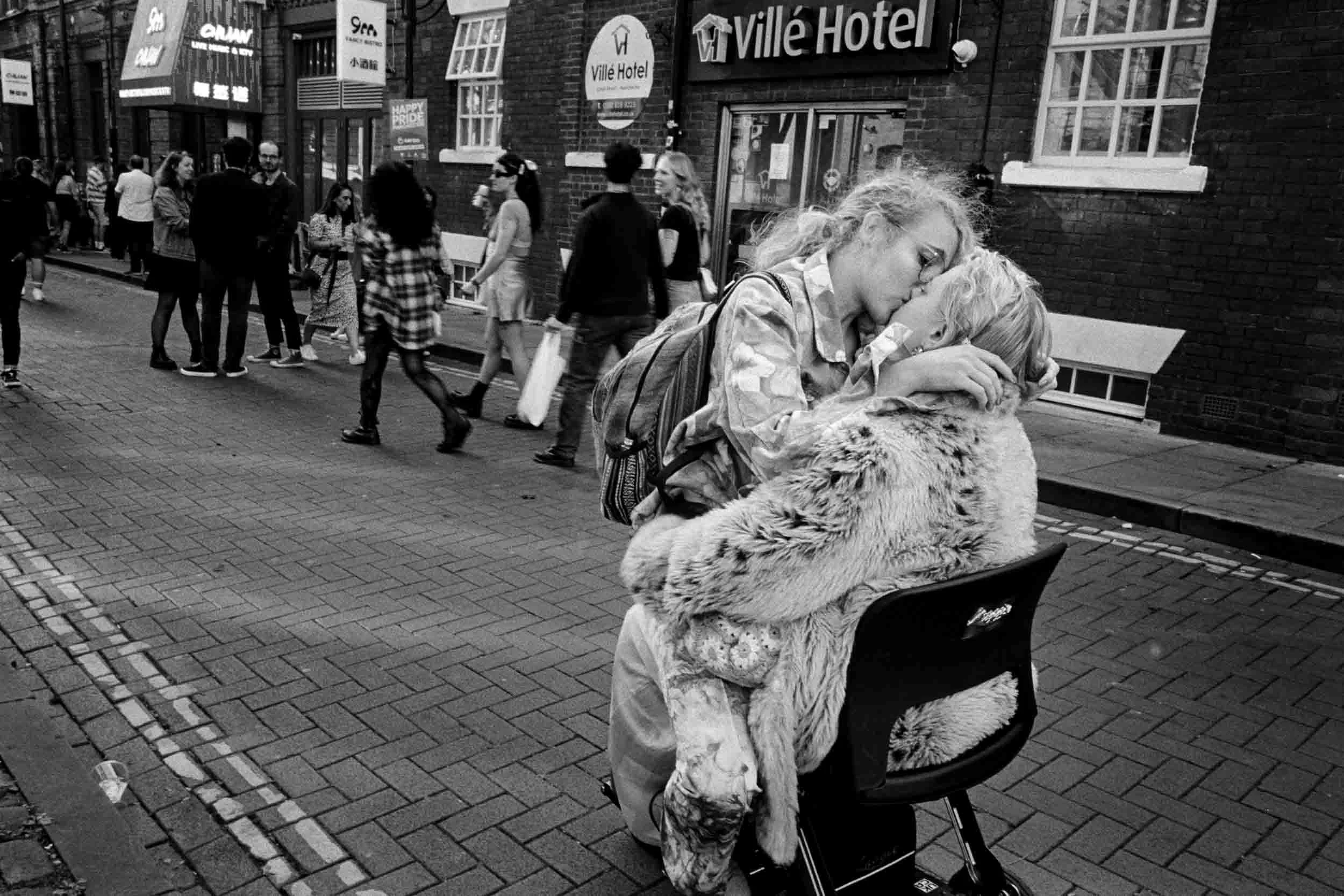 A woman in a furry coat hugging a small dog on a city street near a park bench. Multiple pedestrians walk by, with a hotel and various signs visible on a brick building in the background.
