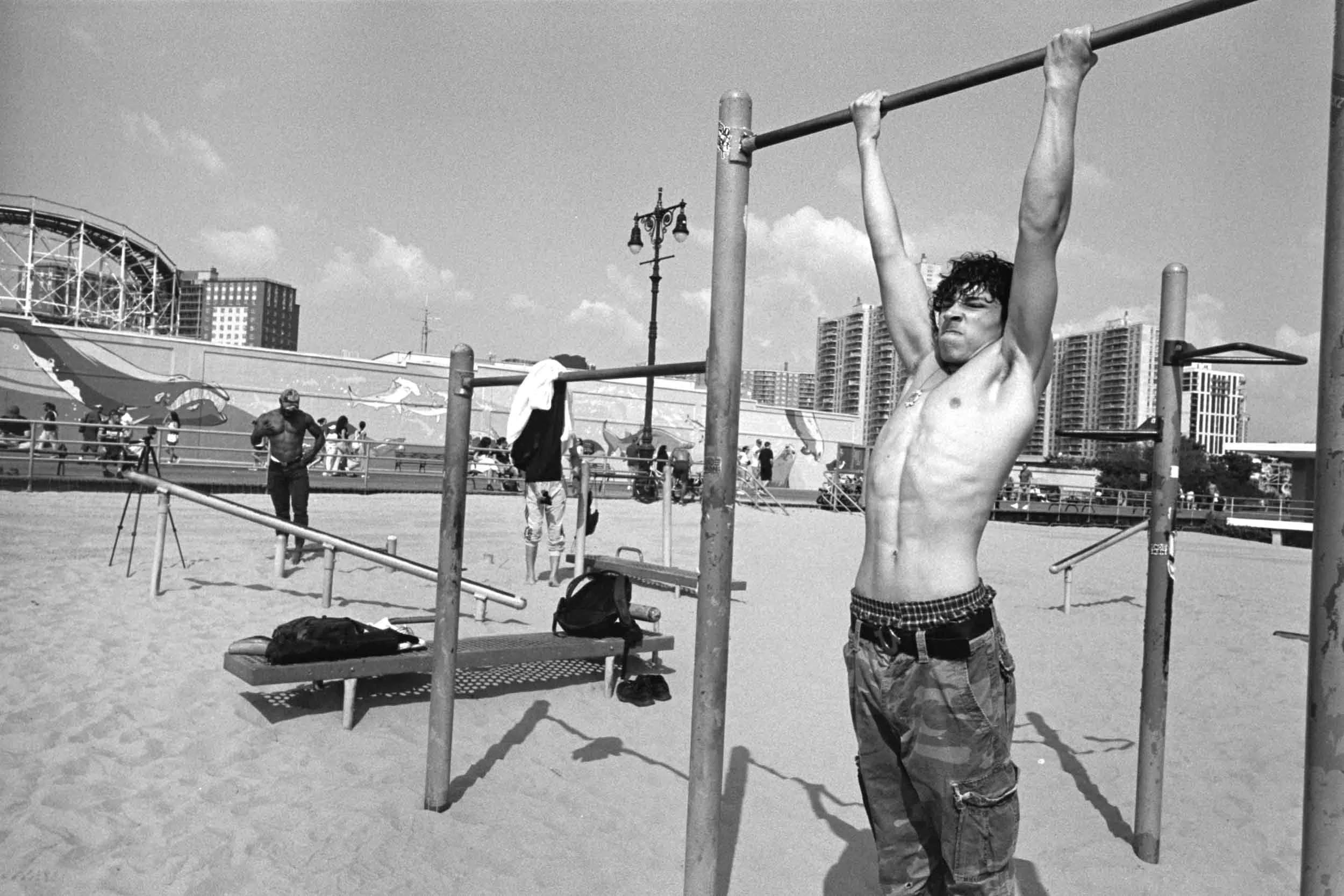 Young man hanging from a pull-up bar in an outdoor gym area at Coney Island with high-rise buildings in the background. Other people are is using the gym and walking around, some standing and stretching. The scene is in black and white.